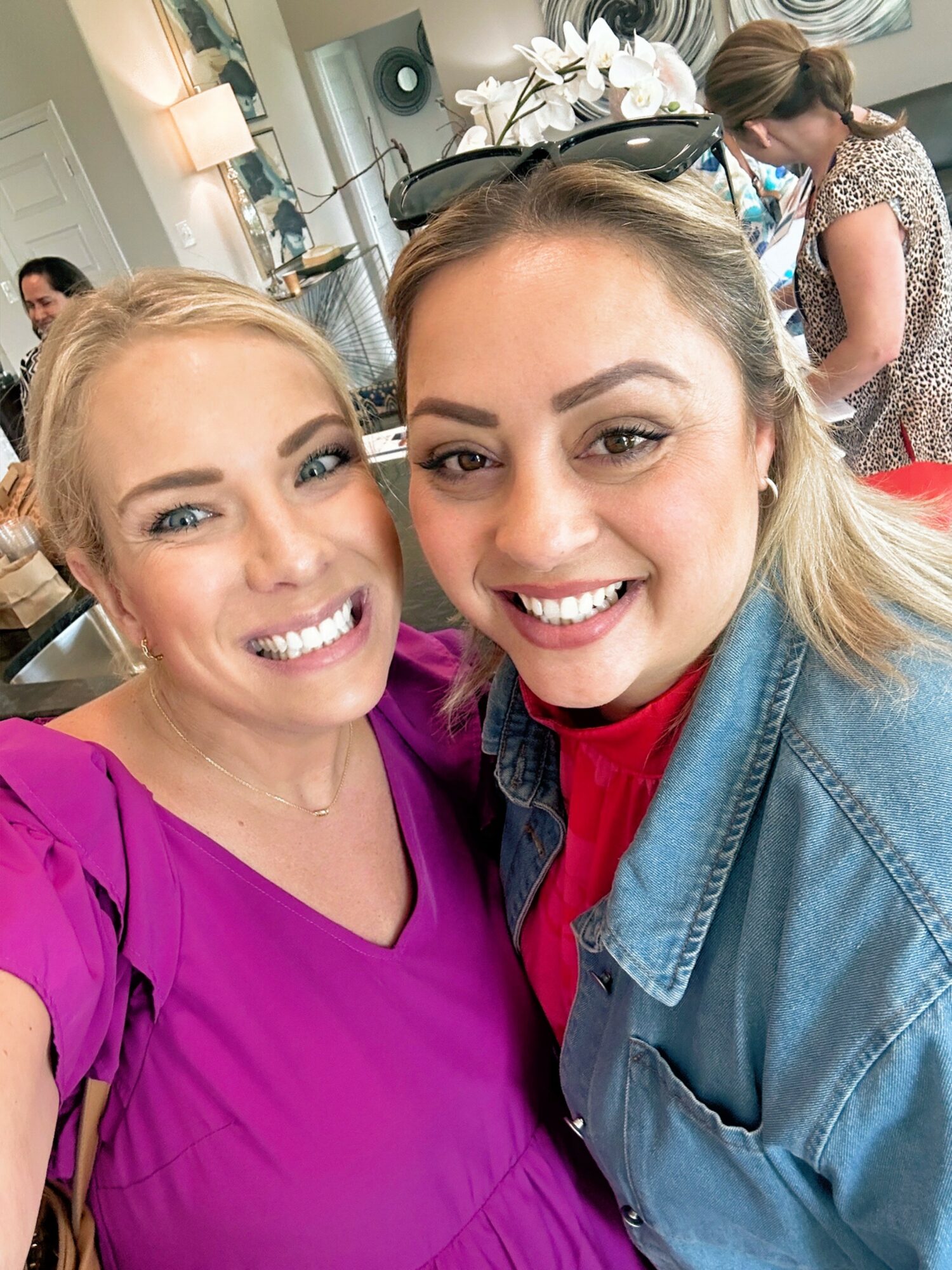 Two women smiling closely, one in a purple top and the other in a denim jacket and red shirt, in a bright indoor setting.