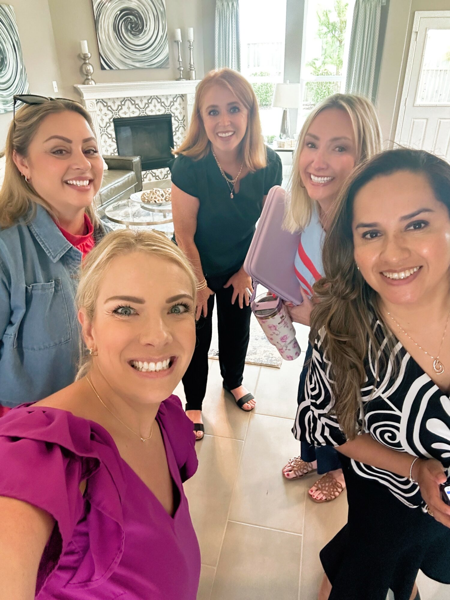 Five women smiling and posing for a photo indoors, with a fireplace and windows in the background.
