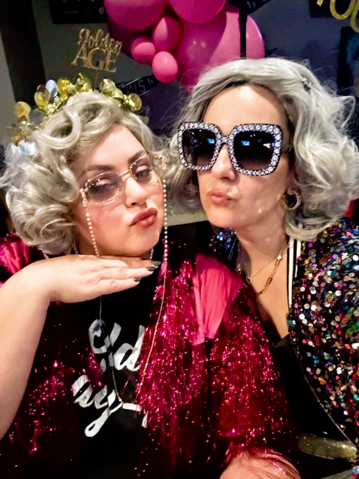 Two women with curly gray hair wearing large sunglasses and colorful outfits, celebrating at a party with pink balloons in the background.