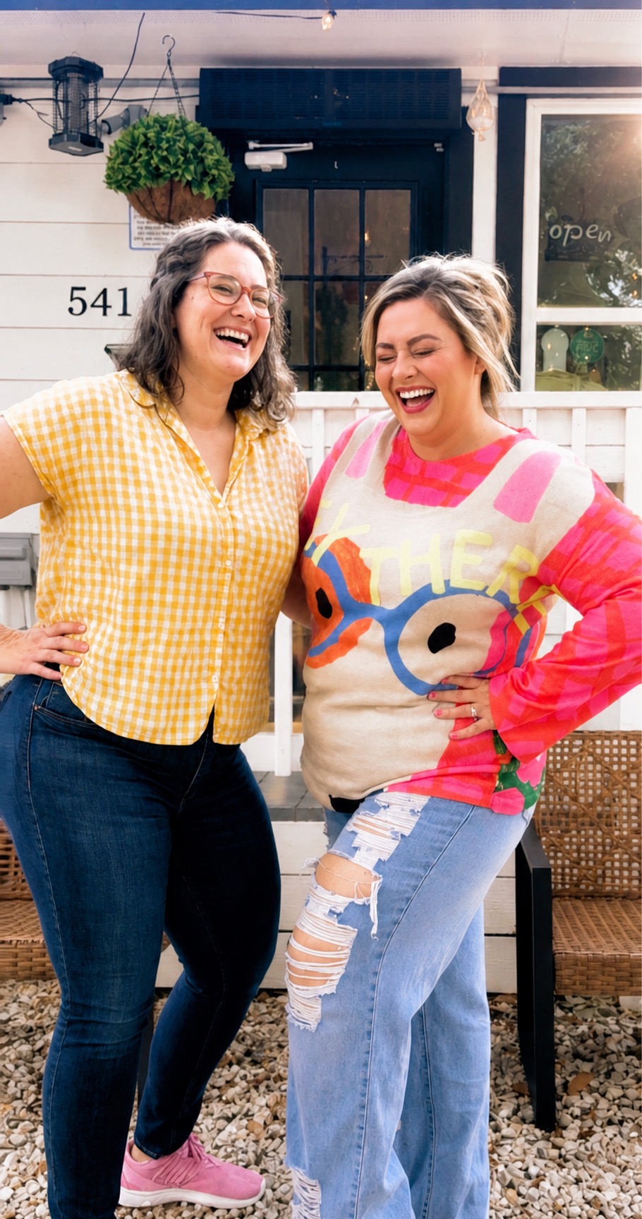 Two women laughing and smiling outside a house, one wearing a yellow checkered shirt, the other in a colorful sweater.