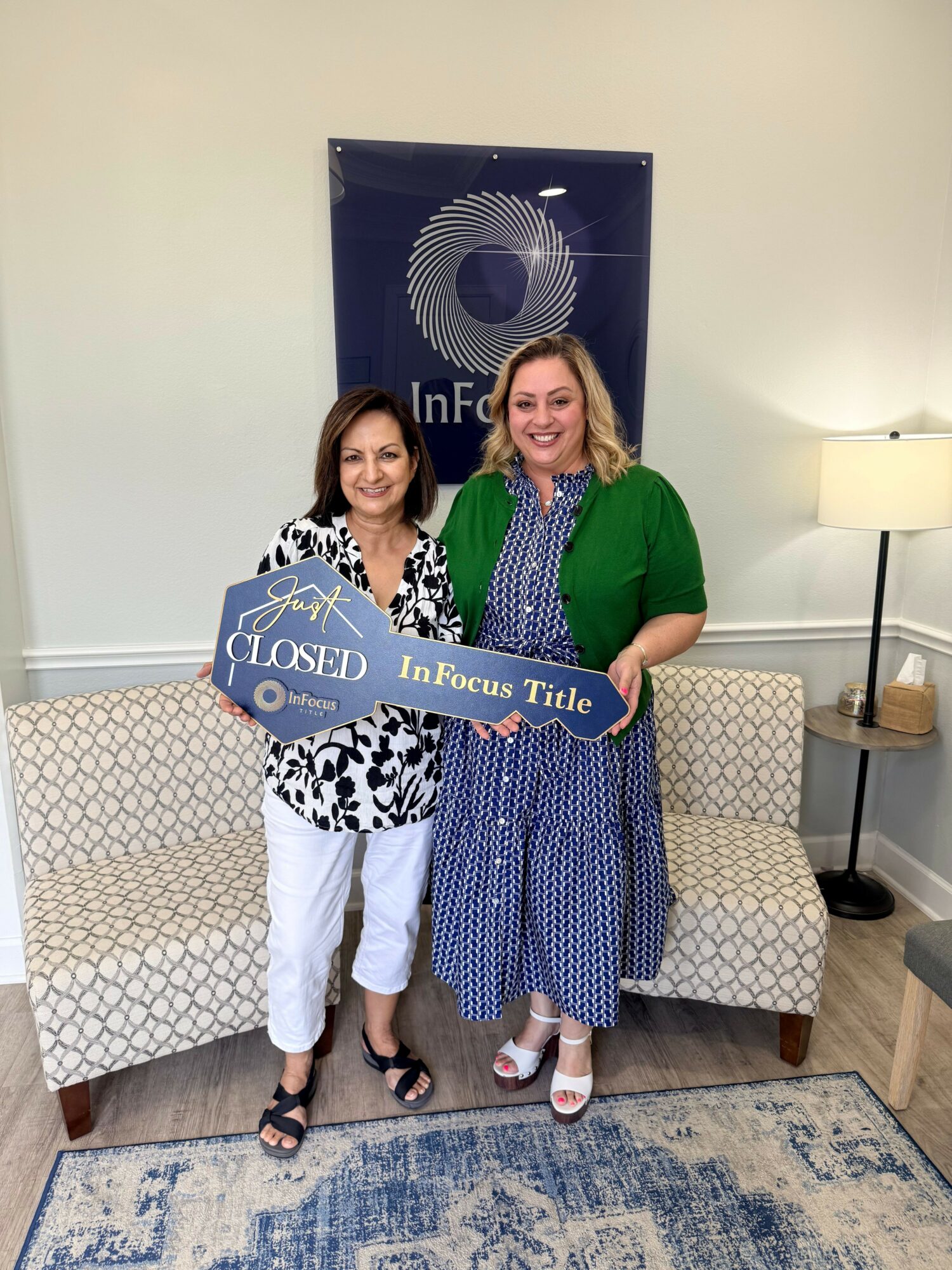 Two women standing indoors, holding a sign that says 'Closed for Lunch,' smiling, with a patterned sofa and a blue and white rug behind them.