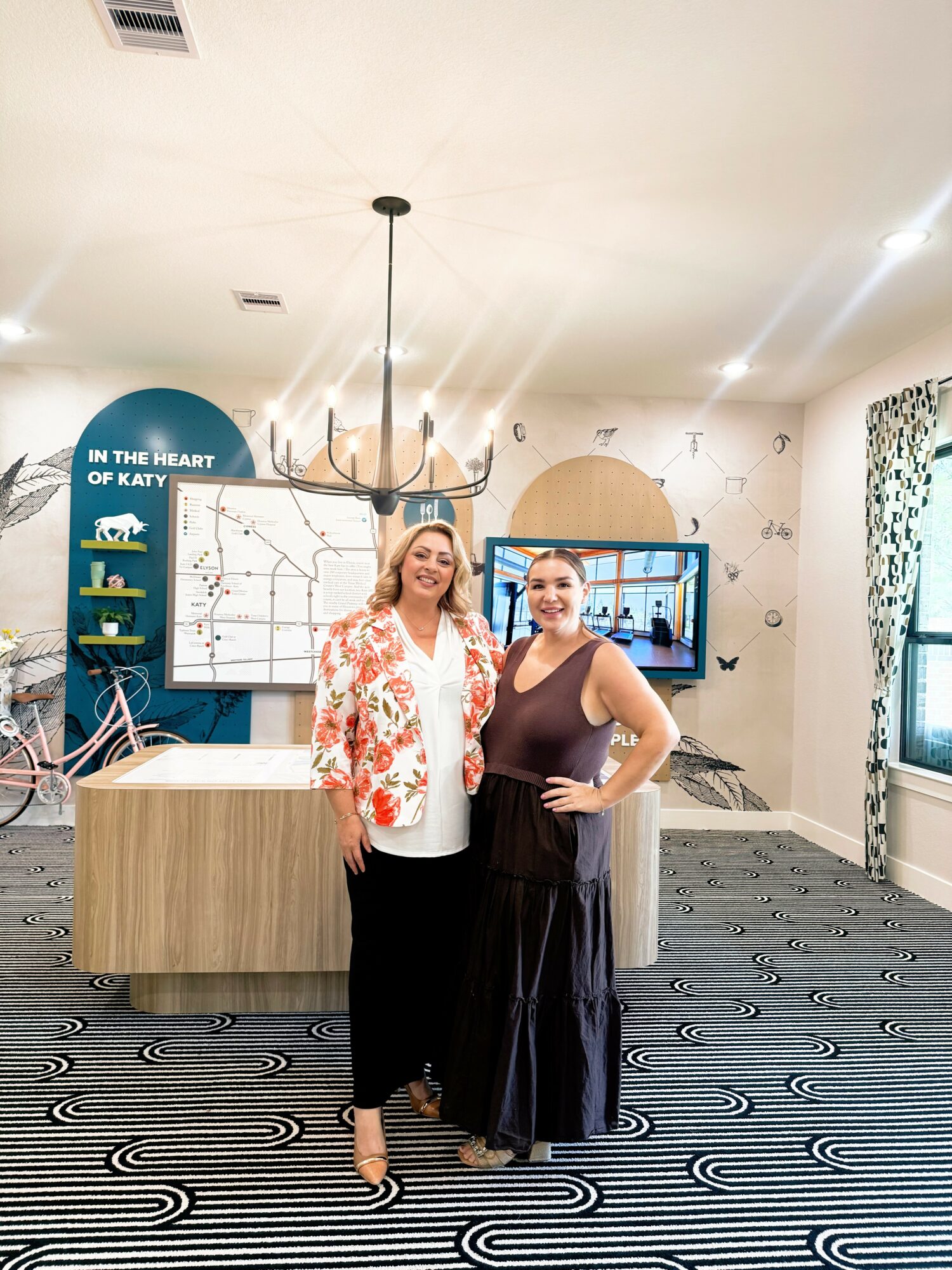 Two women standing in a room with patterned carpet, a chandelier, and a blue wall with shelves and a sign, smiling.