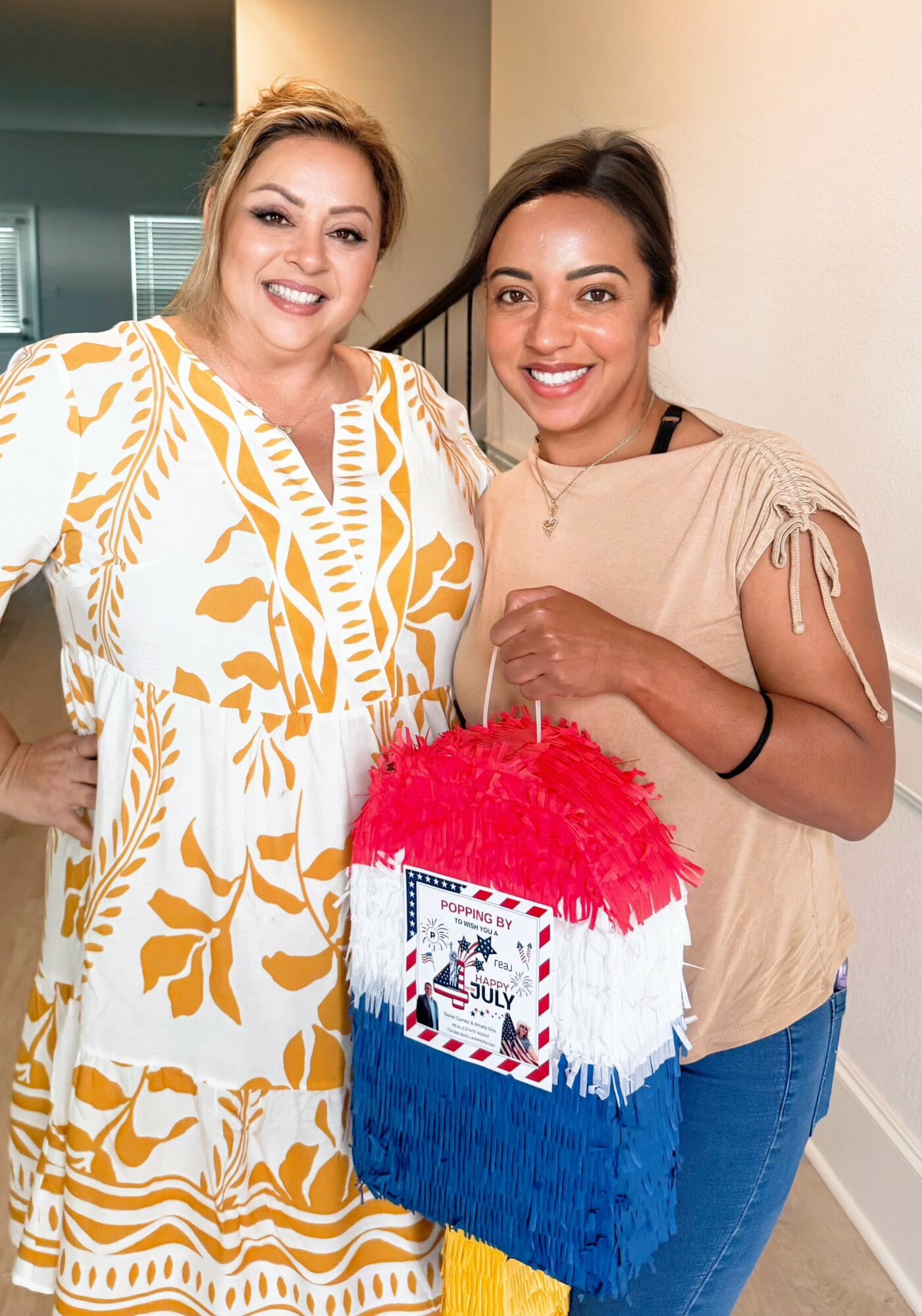 Two women smiling, holding a colorful gift bag with red, white, and blue fringe, indoors with a neutral background.