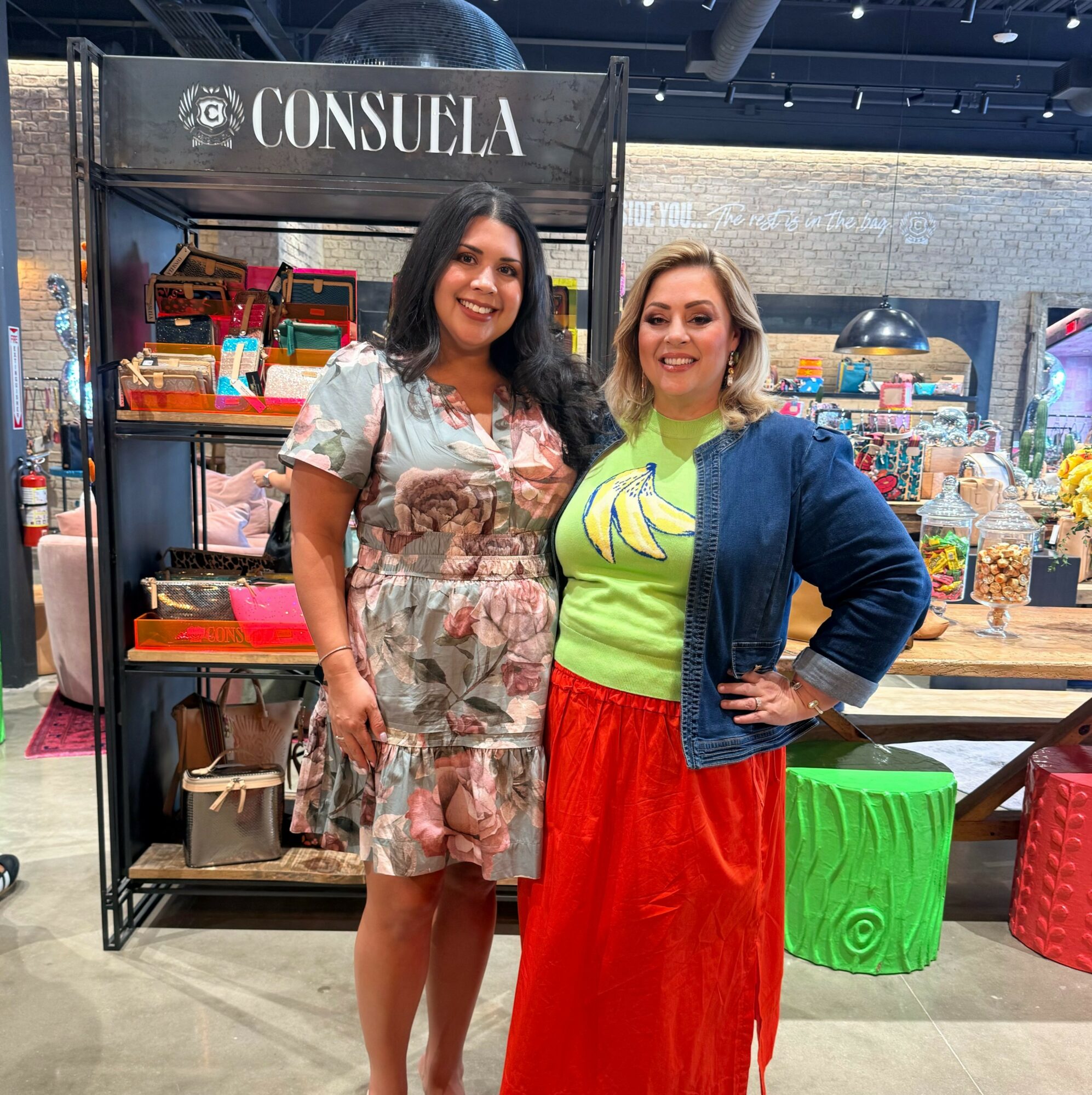 Two women standing in front of a display shelf at a market or store, smiling, with colorful items behind them.
