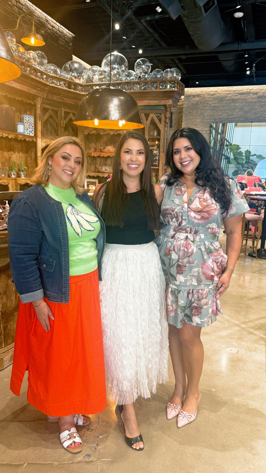 Three women standing together indoors, smiling, with a wooden bar and decorative glass balls overhead in the background.