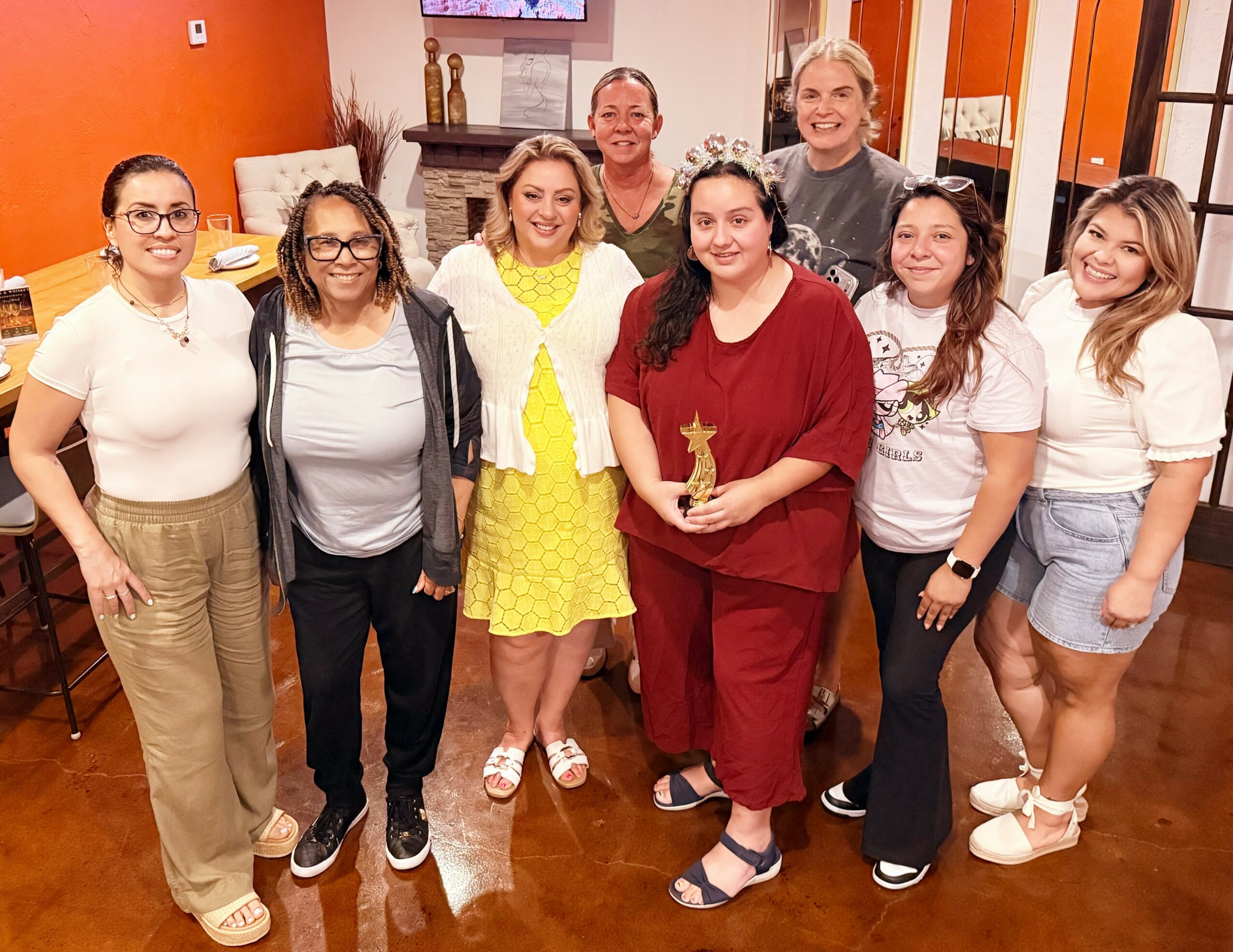 Group of nine women standing indoors, one holding a trophy, smiling, in a warmly decorated room.
