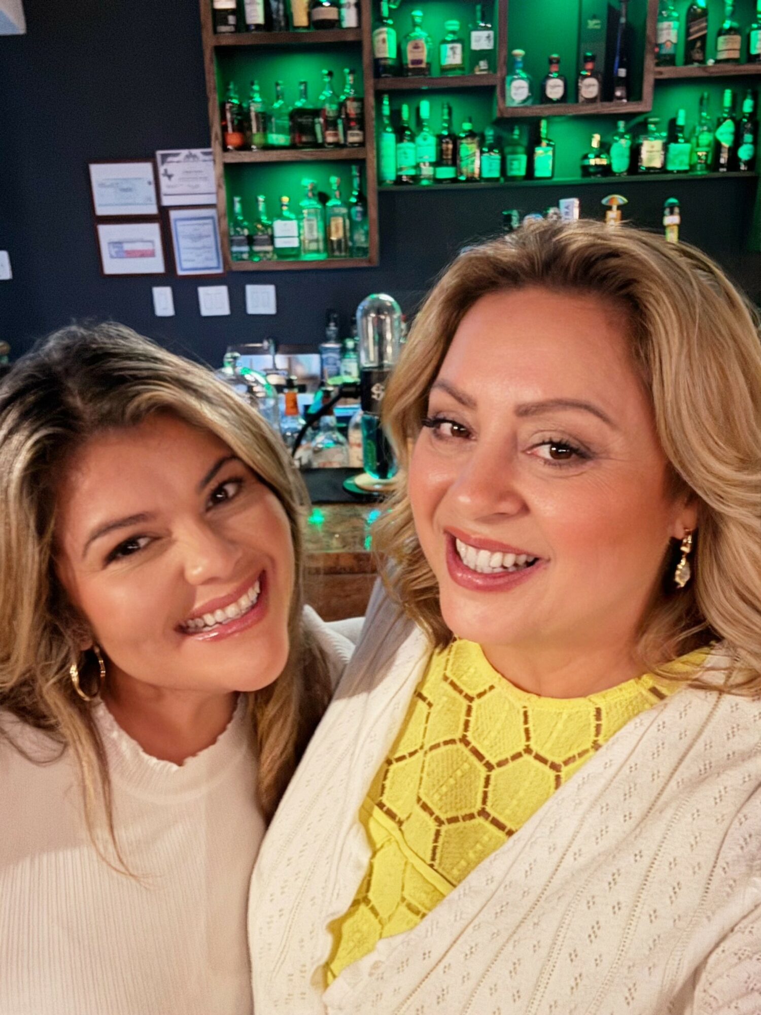 Two women smiling in a bar with green-lit shelves of bottles behind them.