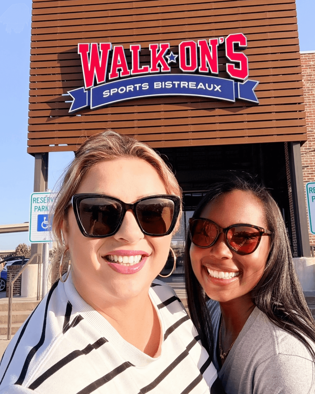 Two women smiling in front of a sign that reads 'Walk-On's Sports Bistreaux' on a sunny day.