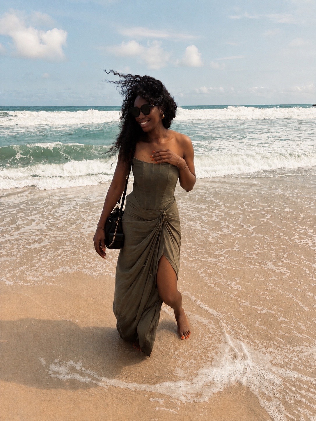 Woman in a strapless dress walking on the beach with waves, holding a bag, hair blowing in the wind.