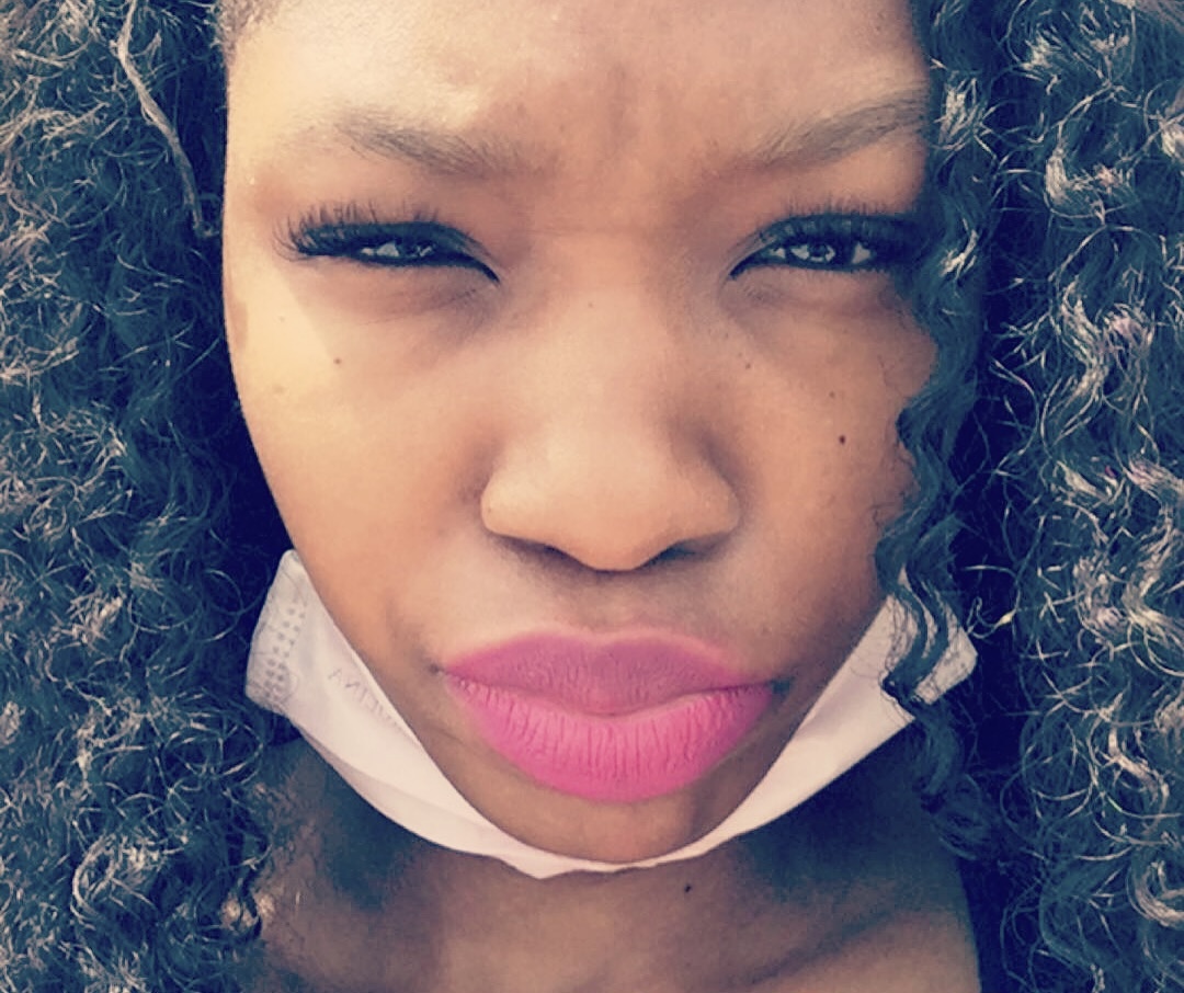 Close-up of a woman with curly hair, squinting eyes, and pink lipstick, wearing a face mask below her chin.