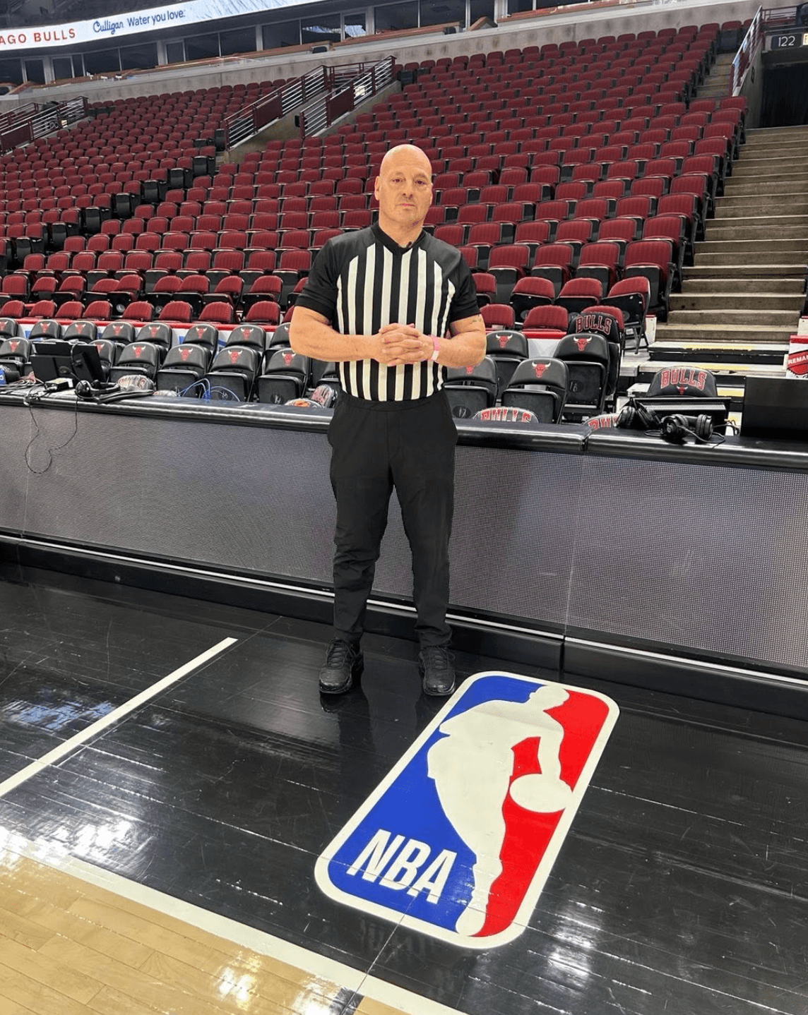 Referee standing on basketball court near NBA logo, empty arena seats in background.