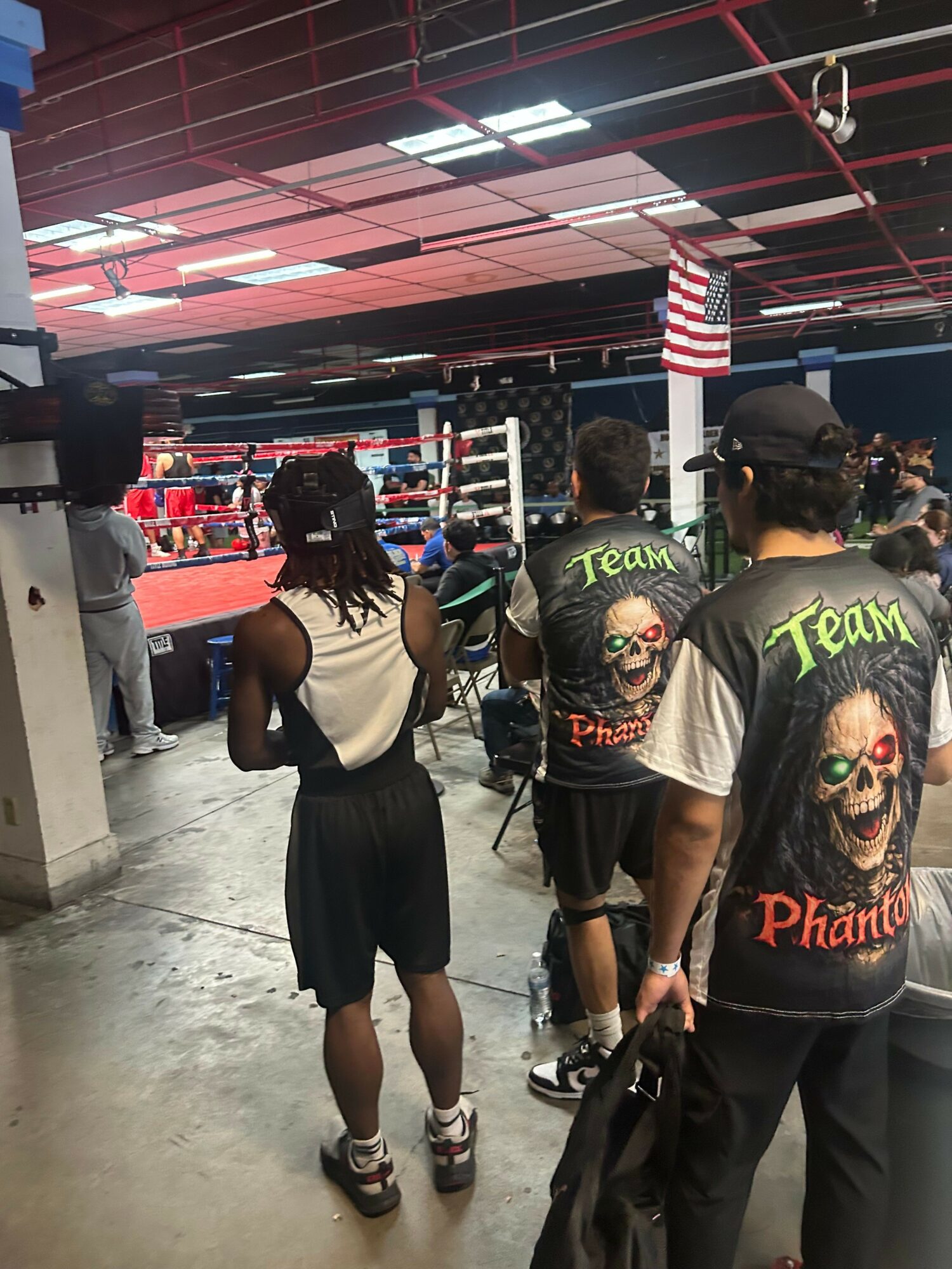 People in a boxing gym with punching bags and boxing ring, some wearing graphic T-shirts, others in casual clothing.
