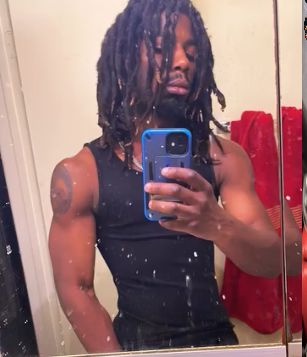 Person with dreadlocks taking a mirror selfie, wearing a black sleeveless shirt, in a room with a red towel in the background.