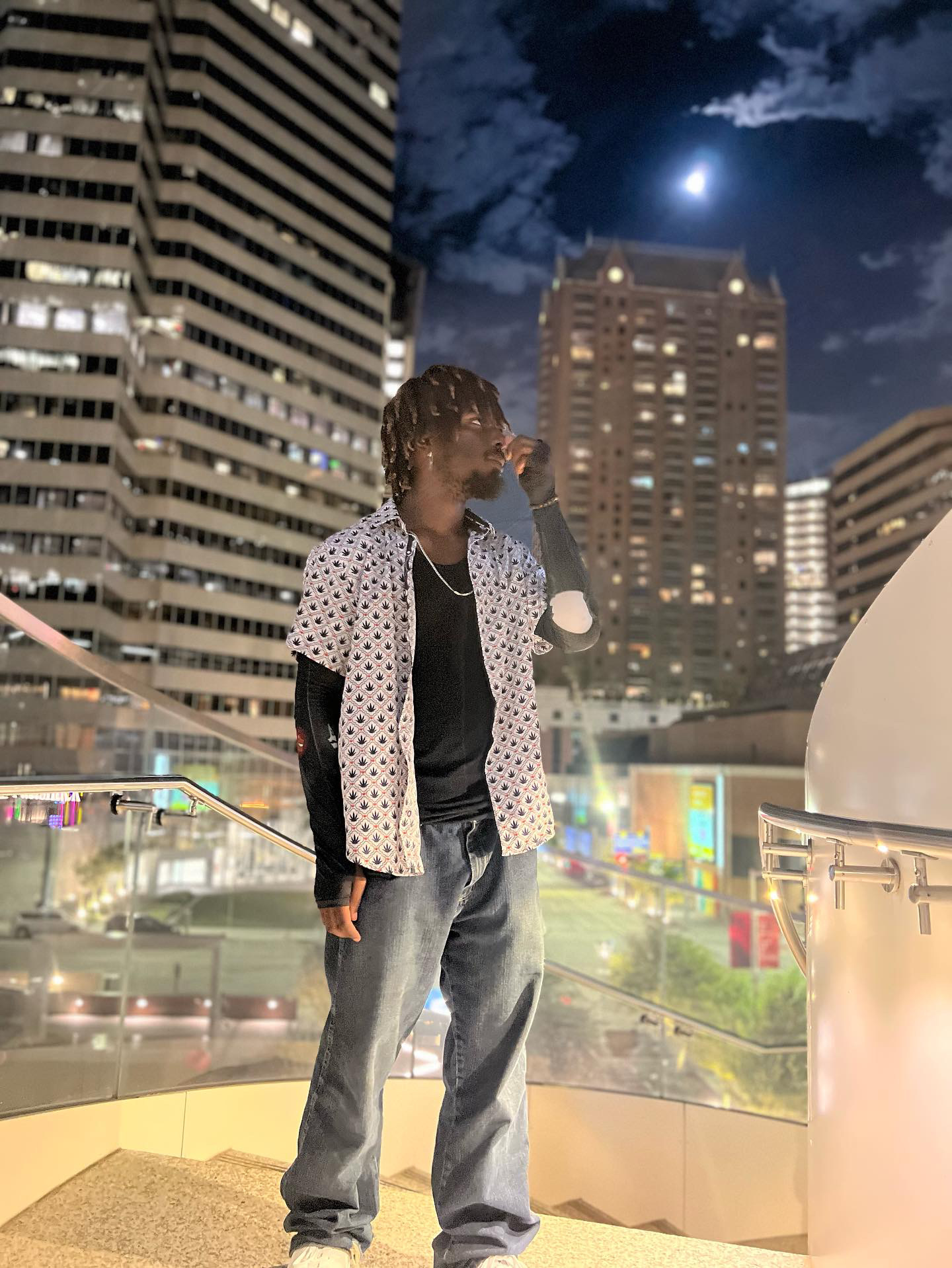Person standing on a balcony at night with city buildings and moon in the sky behind them.