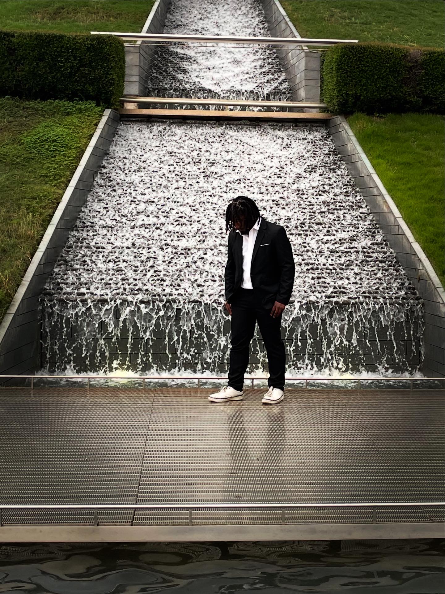 Person in black jacket and white shoes standing in front of a cascading water feature with greenery on sides.