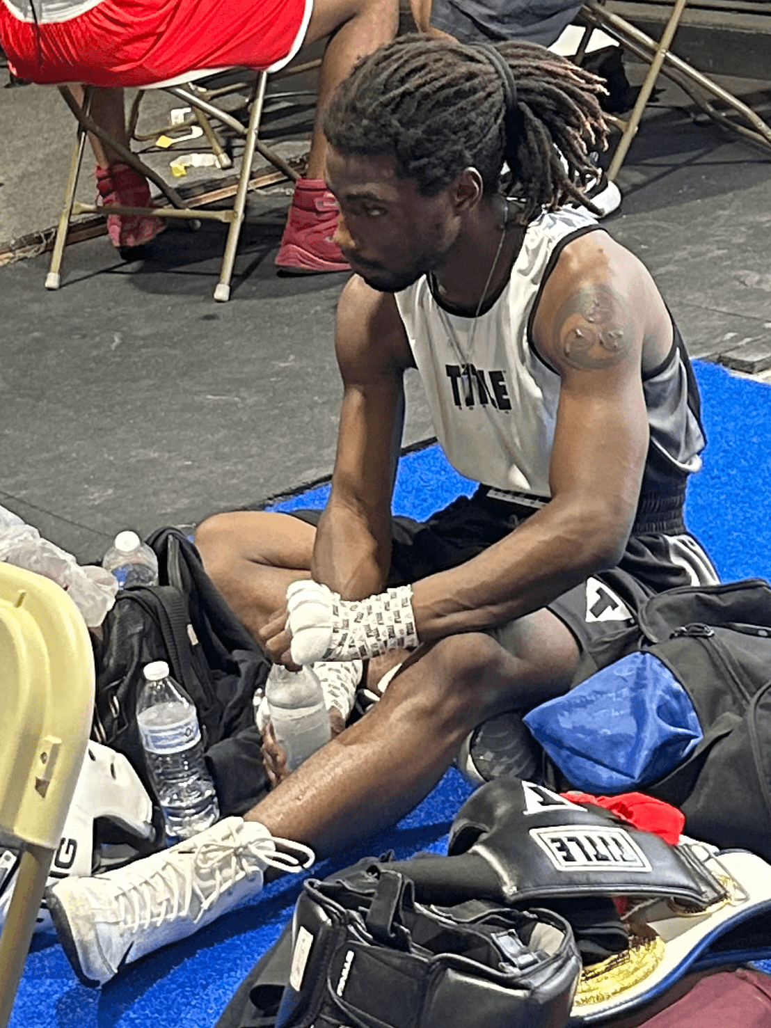Young man with dreadlocks sitting on the floor, wearing a sleeveless shirt, shorts, and white sneakers, surrounded by bags and water bottles.