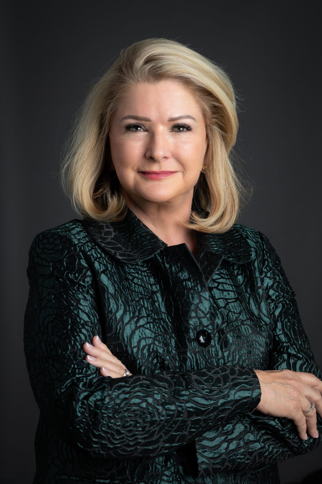 A woman with blonde hair in a dark patterned jacket, arms crossed, smiling slightly against a dark background.