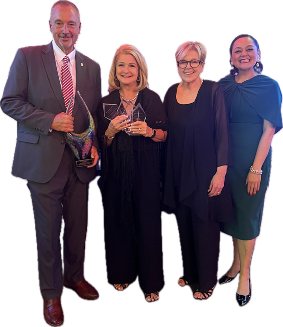 Four people standing together, smiling, with a black background. One man and three women, dressed formally, holding an award.