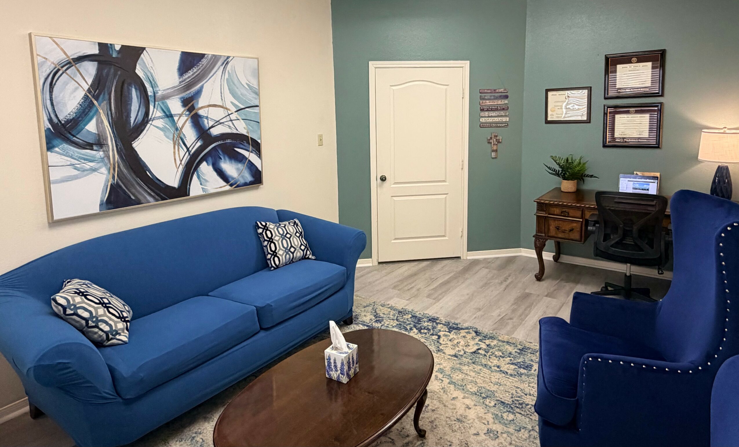 Living room with blue sofa, armchair, abstract painting, and a wooden desk with framed pictures on green wall.