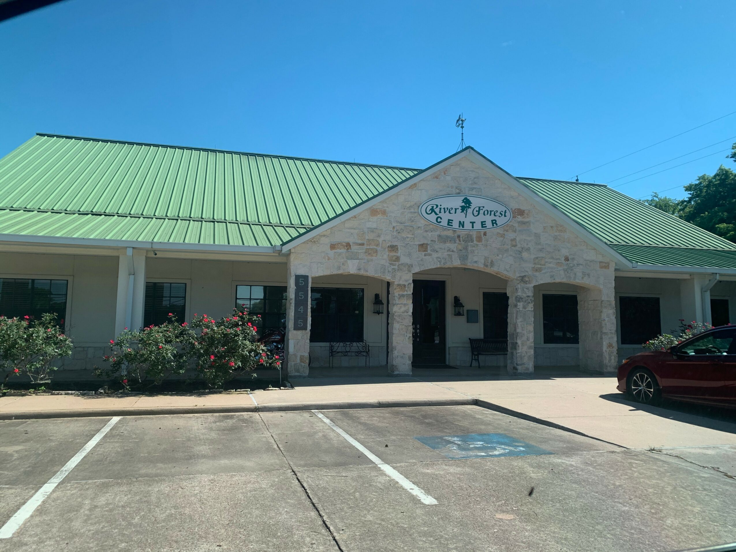 Building with a green roof and stone facade, sign reads 'River of Life Center,' parking lot in front, blue sky above.