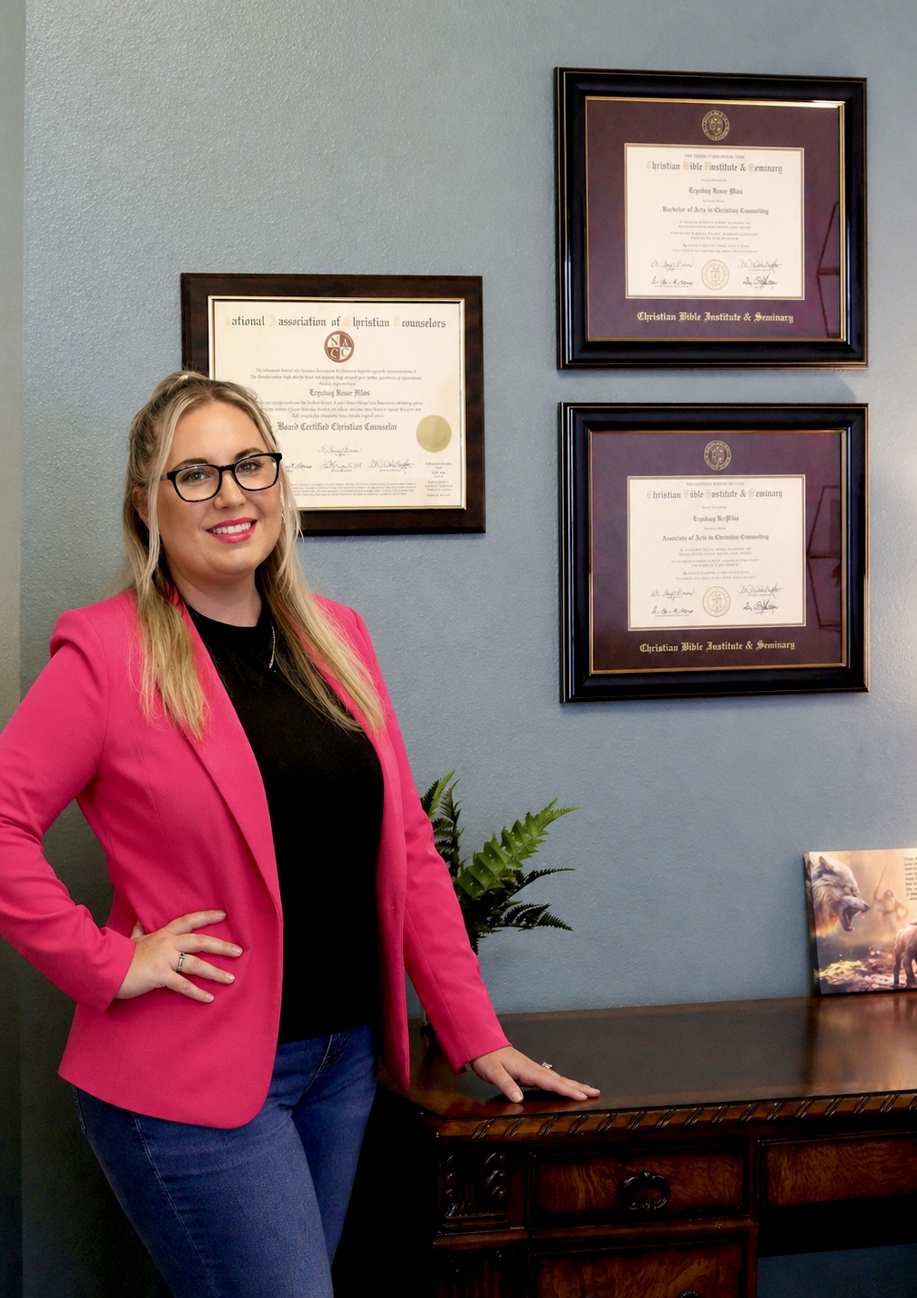 Woman with glasses and blonde hair, wearing a pink blazer and black top, standing next to framed diplomas on a blue wall.
