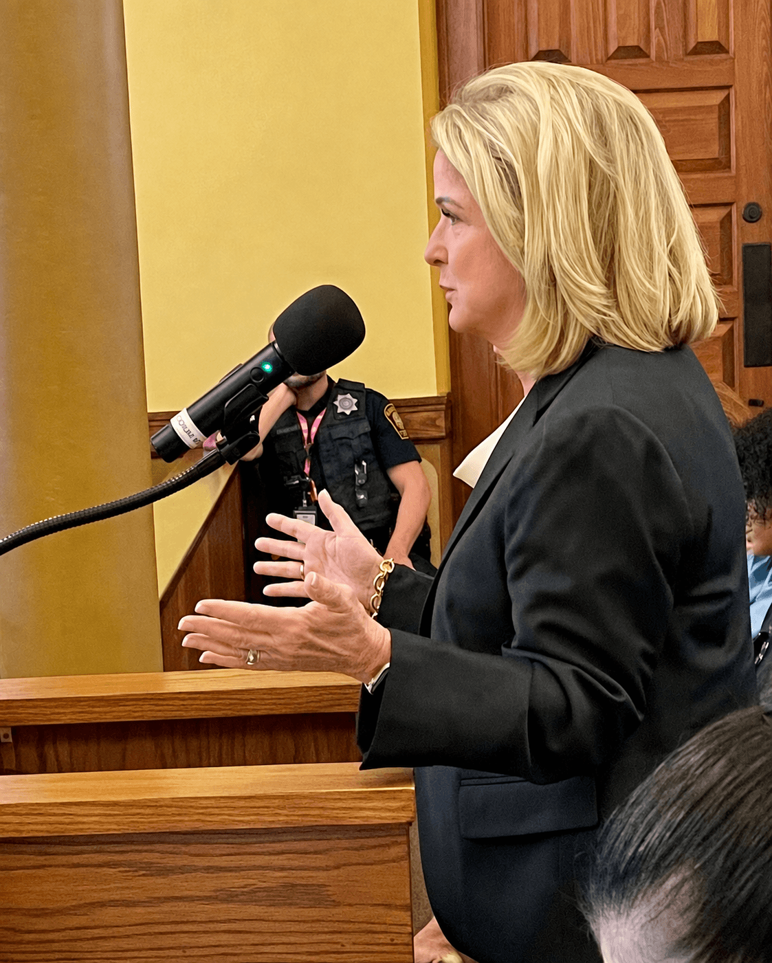 Woman speaking into a microphone, gesturing with hands, in a formal setting with wooden paneling.