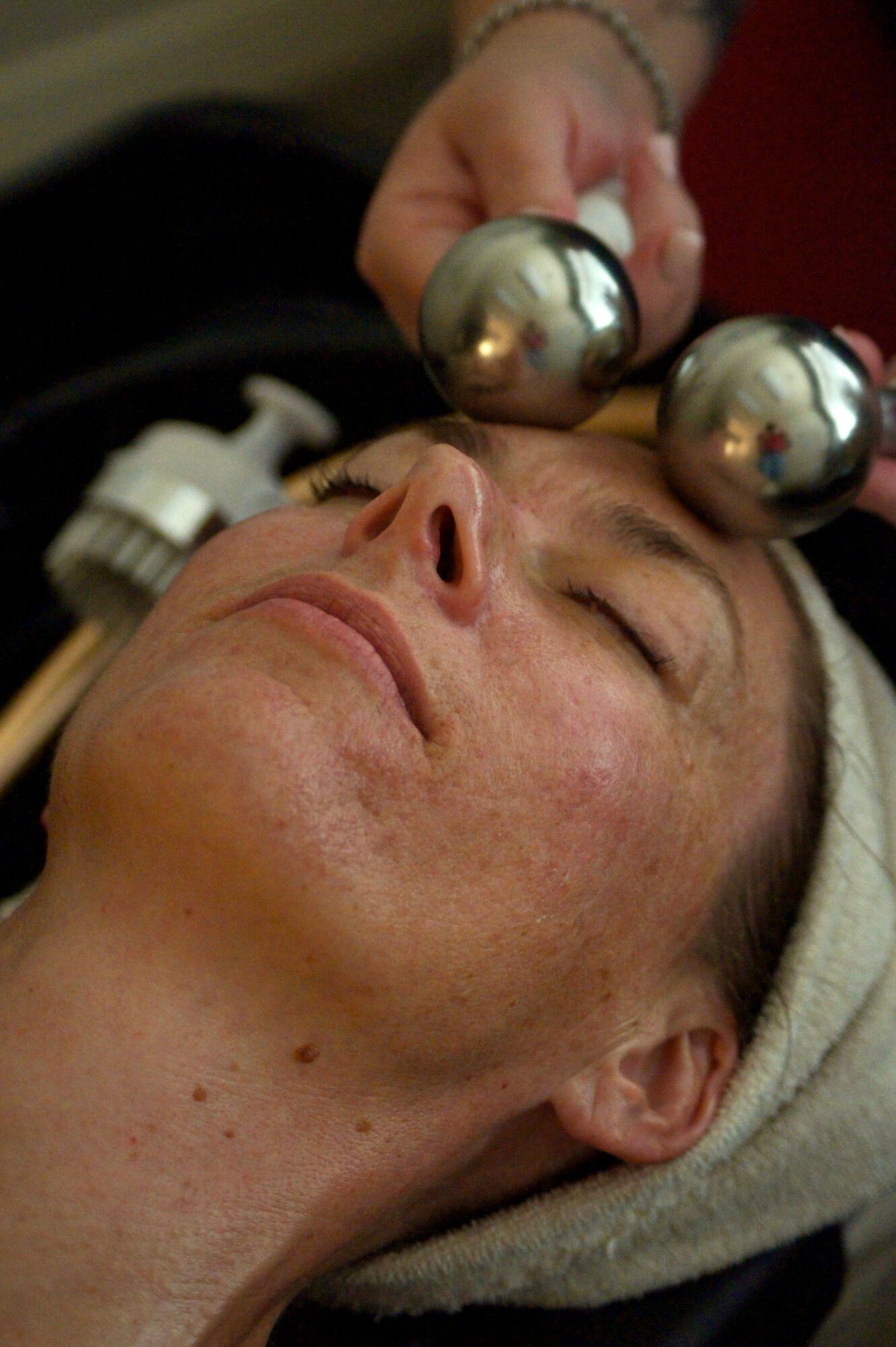 Person receiving a facial treatment with metal tools on forehead, lying down with eyes closed, head wrapped in a towel.