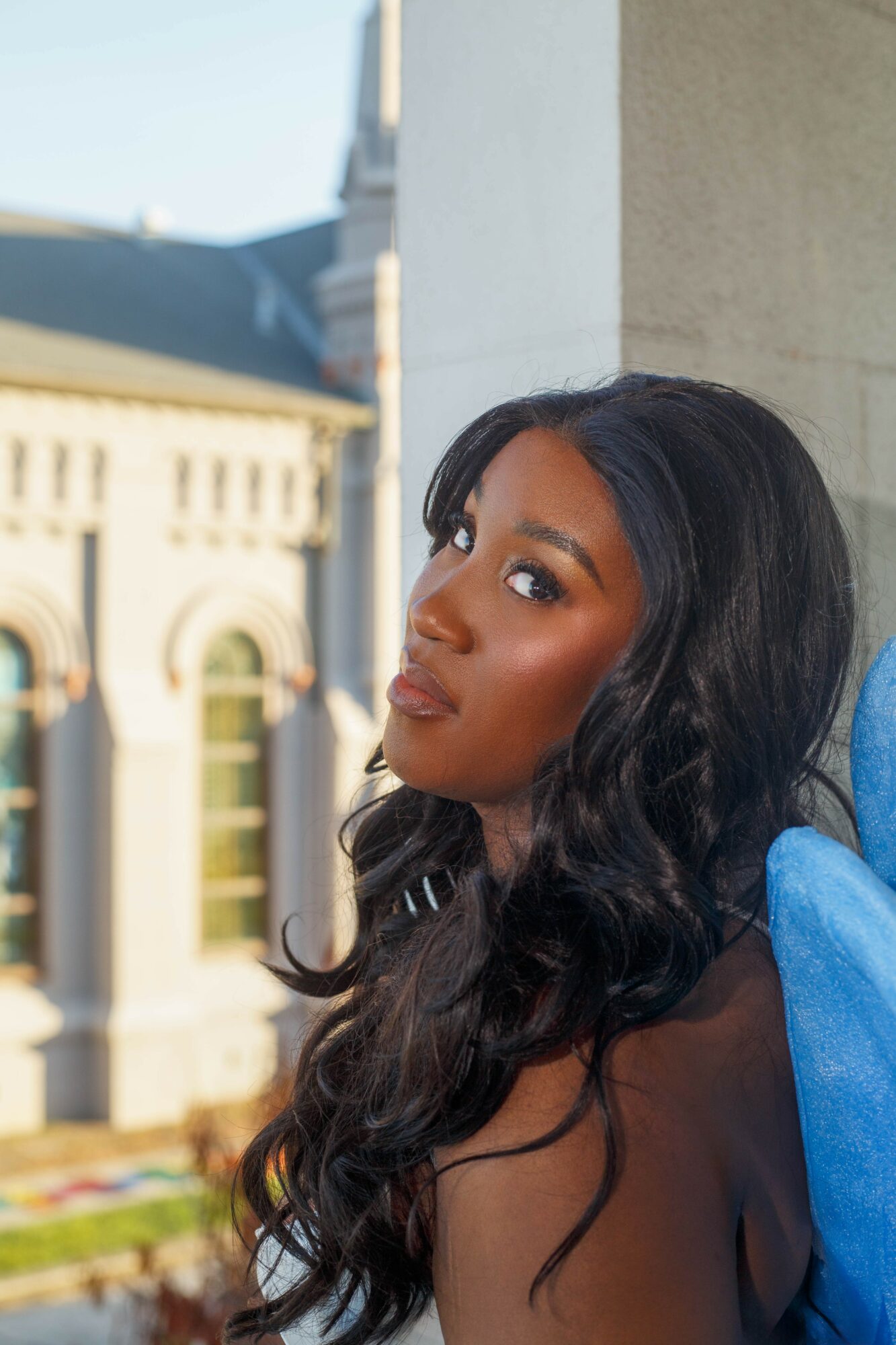 Young woman with long wavy hair looking at camera outdoors, city buildings in background.