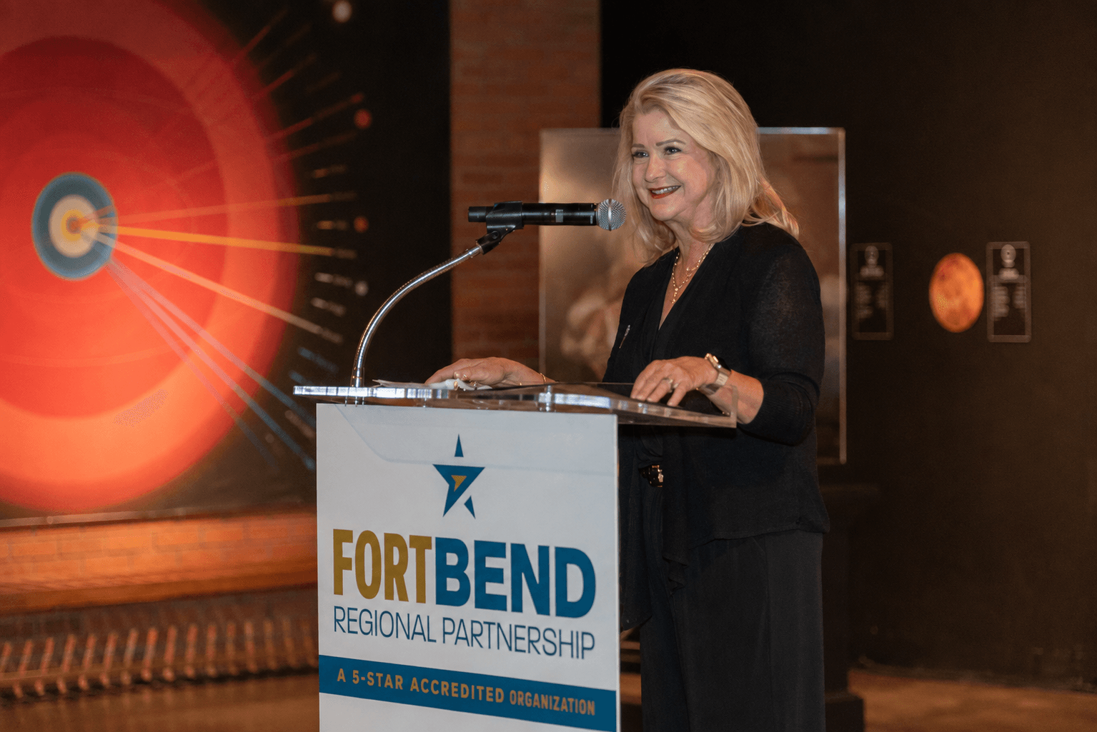 Woman speaking at a podium with a sign reading 'Fort Bend Regional Partnership,' smiling, in a dark room with artwork in the background.