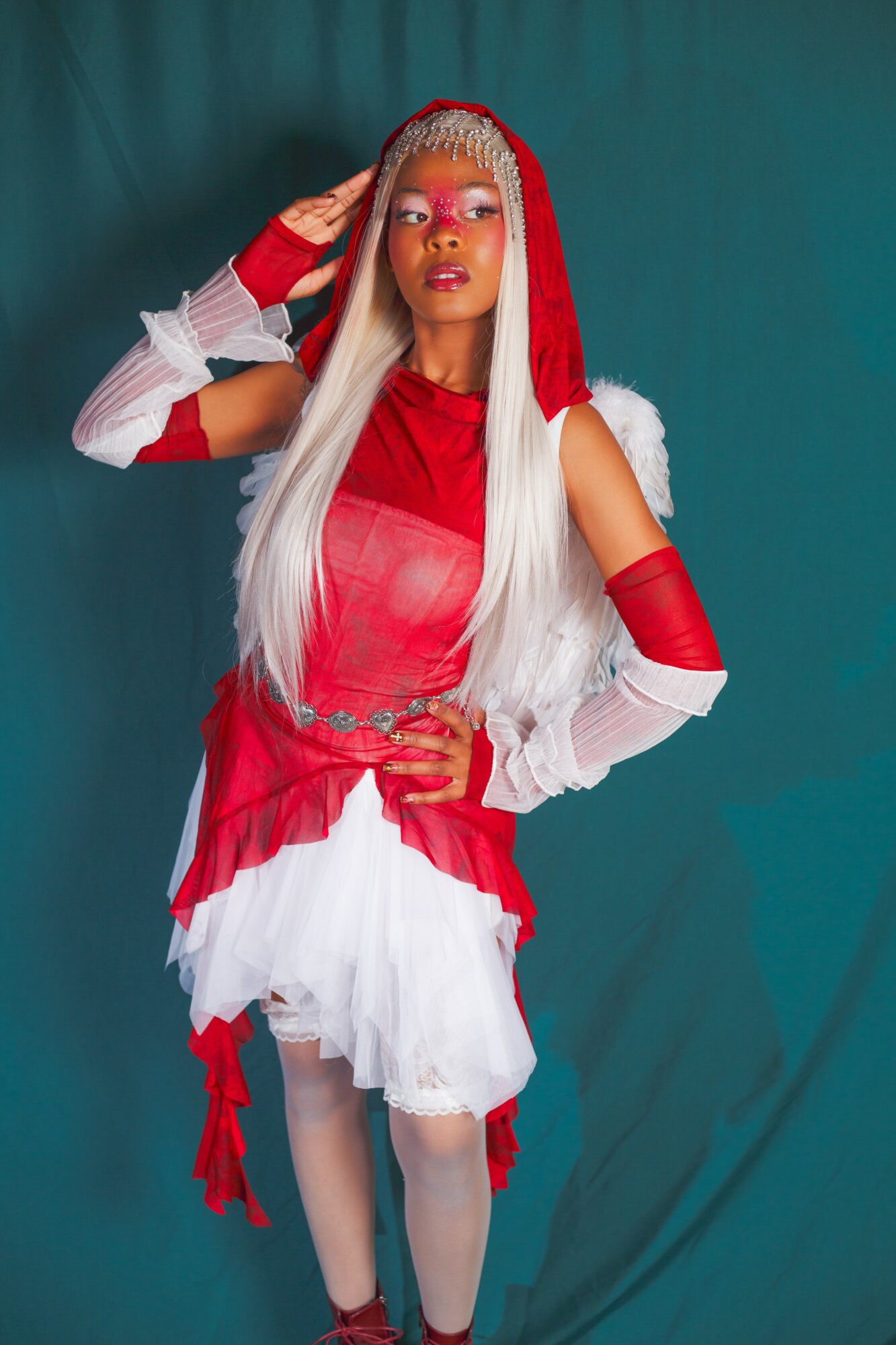 Woman with long white hair wearing red and white costume, posing with hand on head, against teal background.