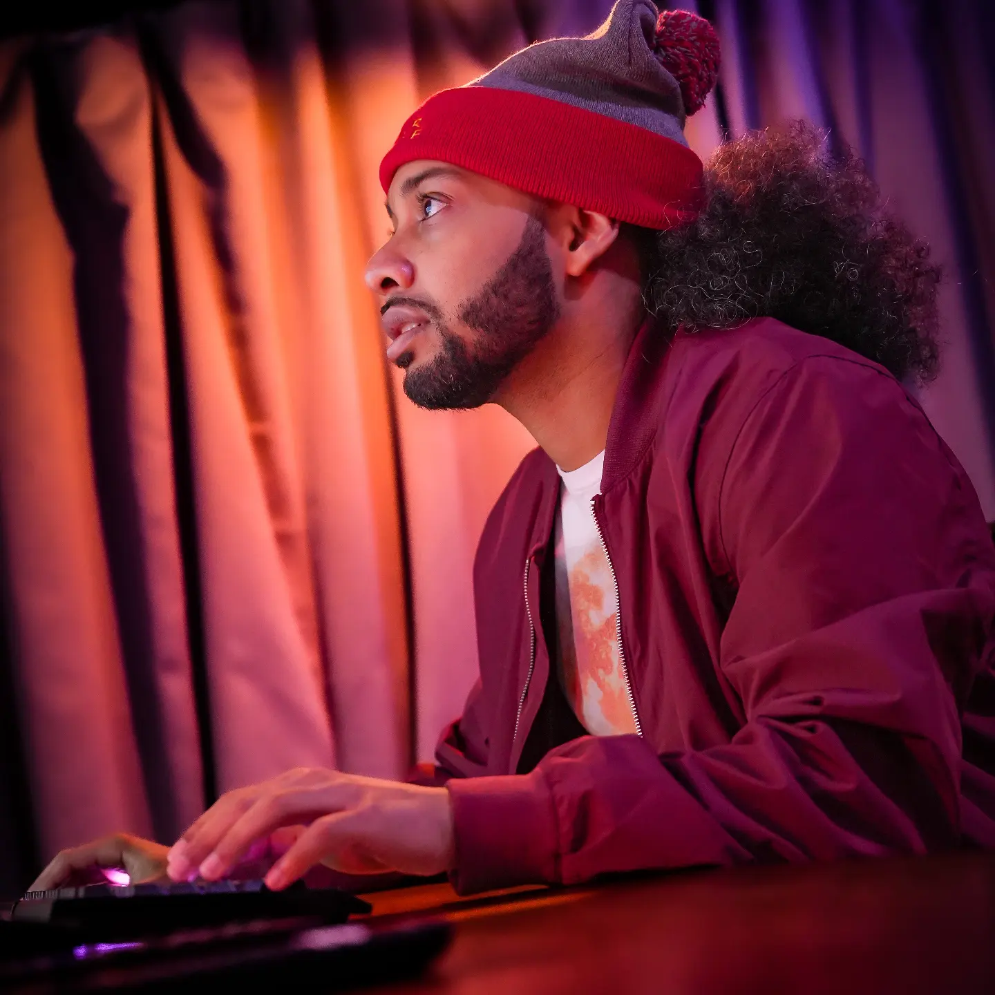 Man with a beard and curly hair wearing a red beanie and maroon jacket, sitting at a table with colorful curtains behind him.