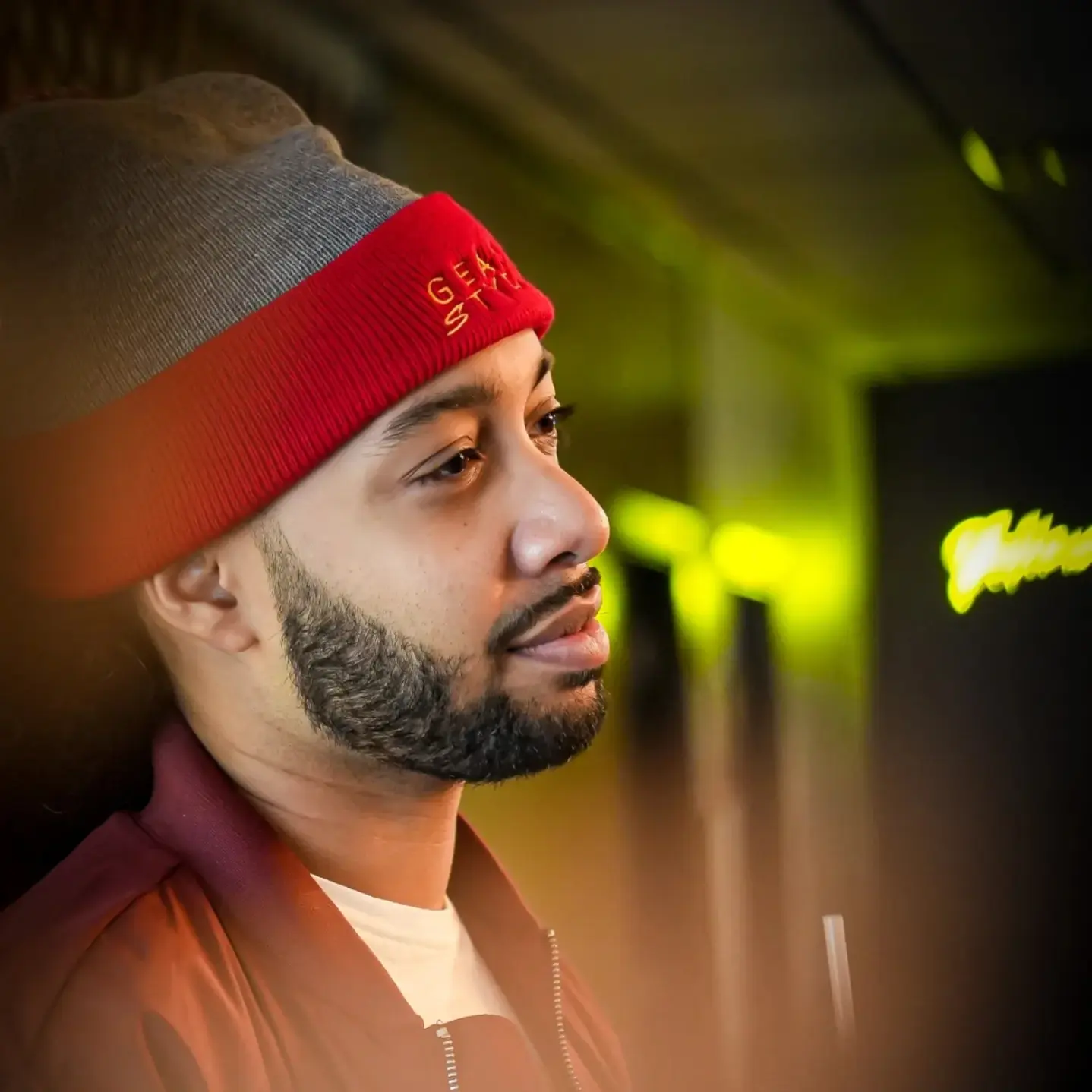 Side profile of a man wearing a gray beanie with red band, beard, and casual clothing, in a dimly lit environment.