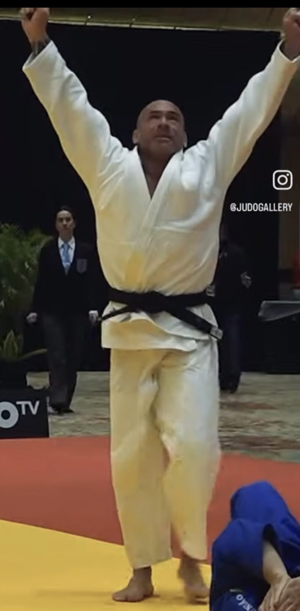 Judo athlete in white gi with black belt raising arms in victory, standing on mat, with referee and audience in background.