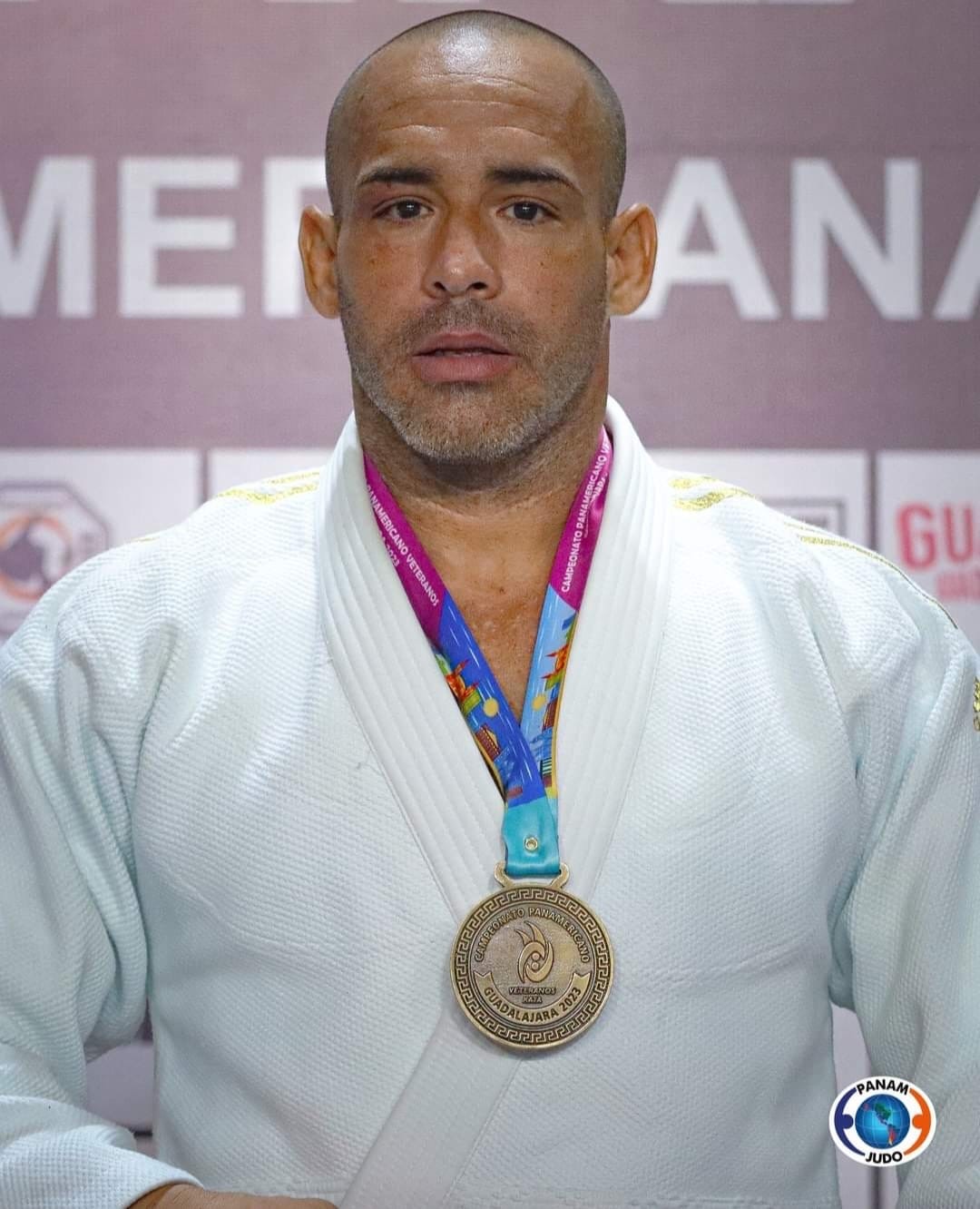 A man in a white judo gi with a medal around his neck, standing in front of a purple background.
