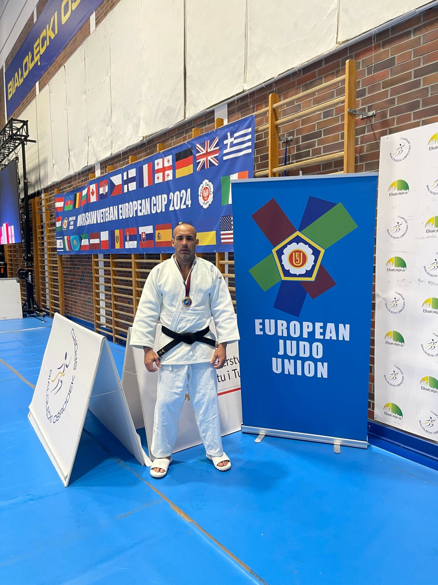 Person in martial arts uniform standing on blue mat, holding a black belt, with banners and flags in background.