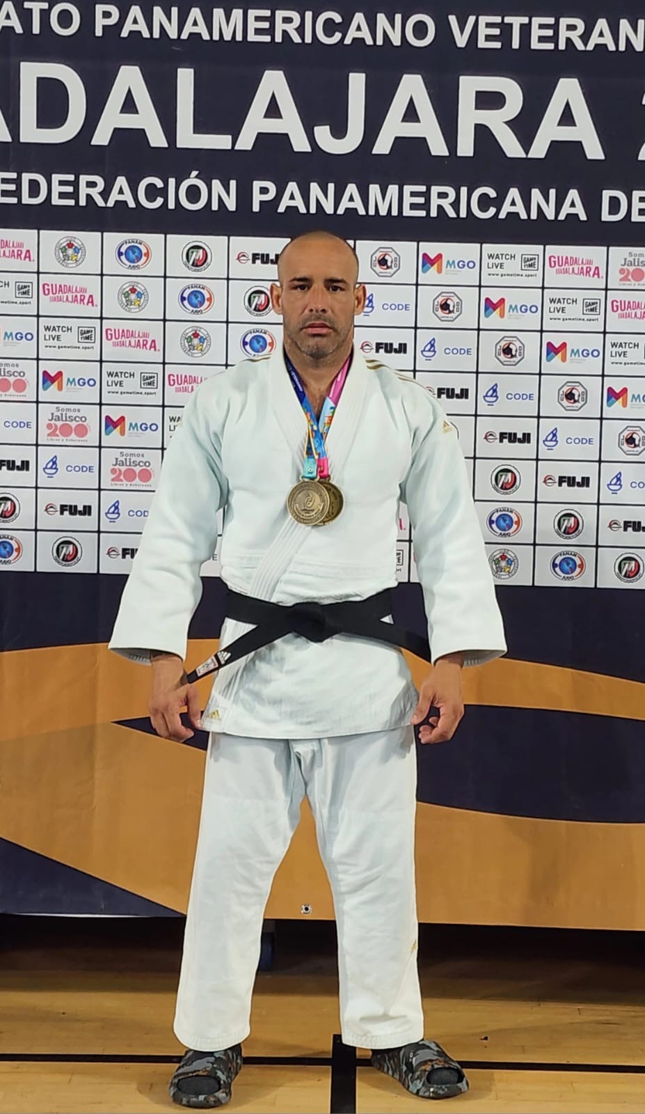 Male judo athlete in white gi with black belt and medal, standing in front of a banner with logos and text.
