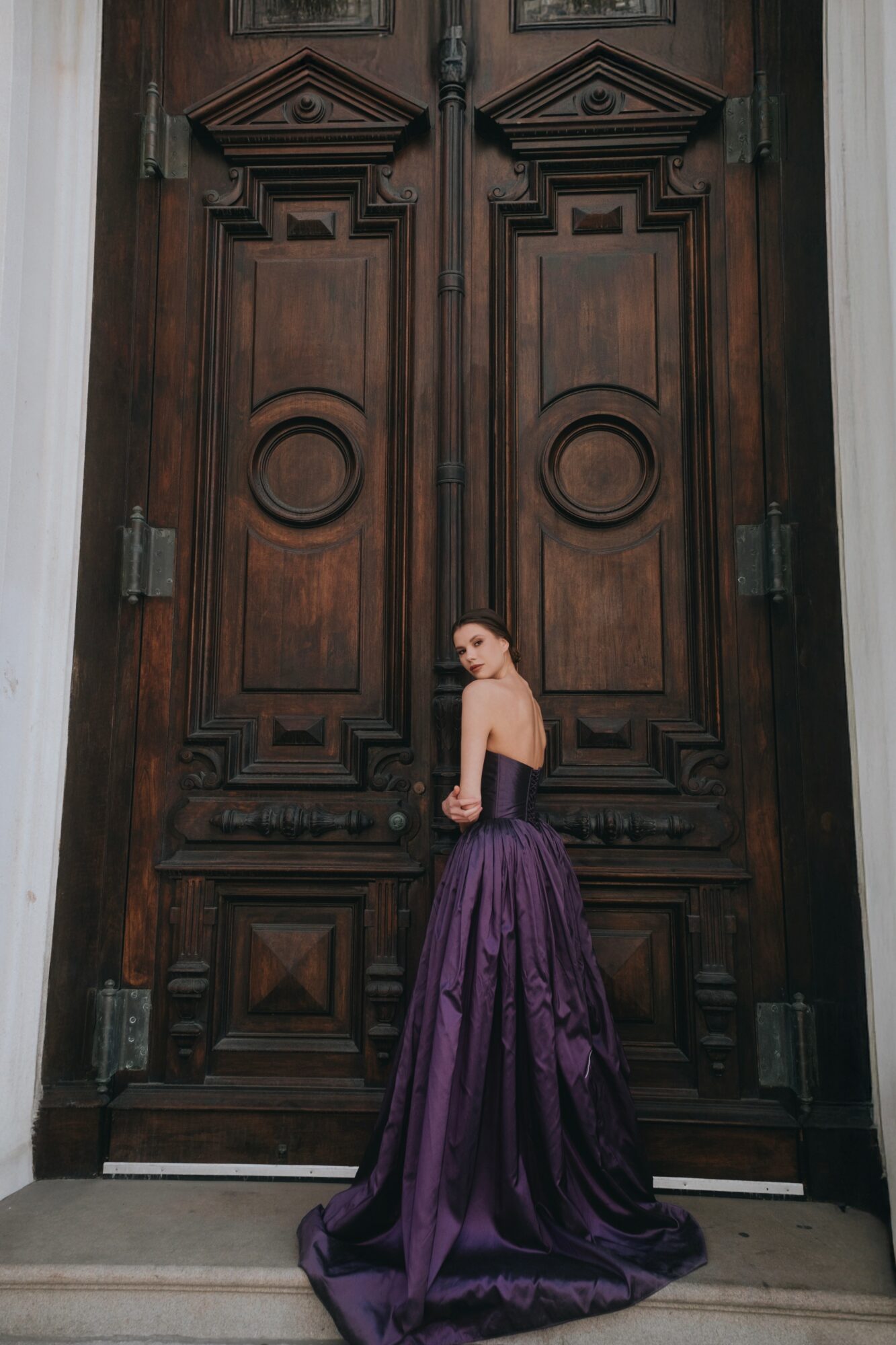 Woman in purple gown standing in front of large wooden door with intricate carvings.