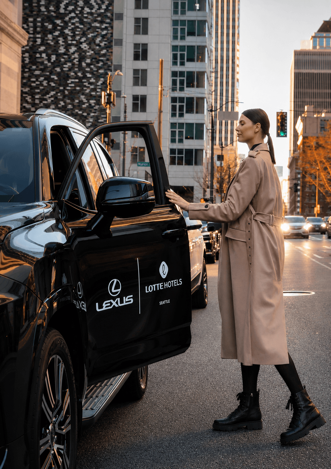 Woman in beige coat and black boots standing outside a black Lexus vehicle in an urban setting with tall buildings.