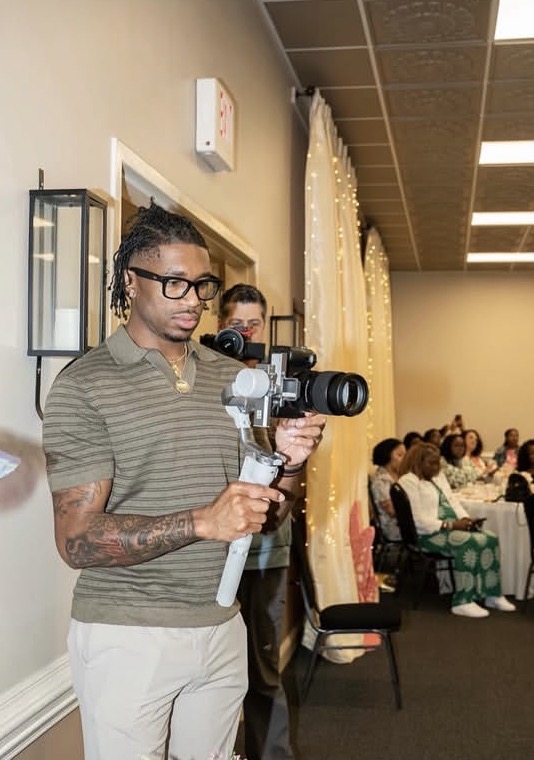 Person with glasses and tattoos holding a camera stabilizer in a room with seated audience, beige walls, and curtains.