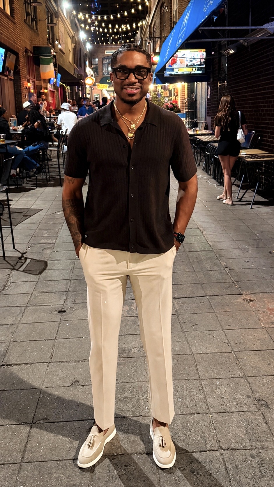 Man standing on a lively city street at night, wearing sunglasses, a dark shirt, light pants, and sneakers.