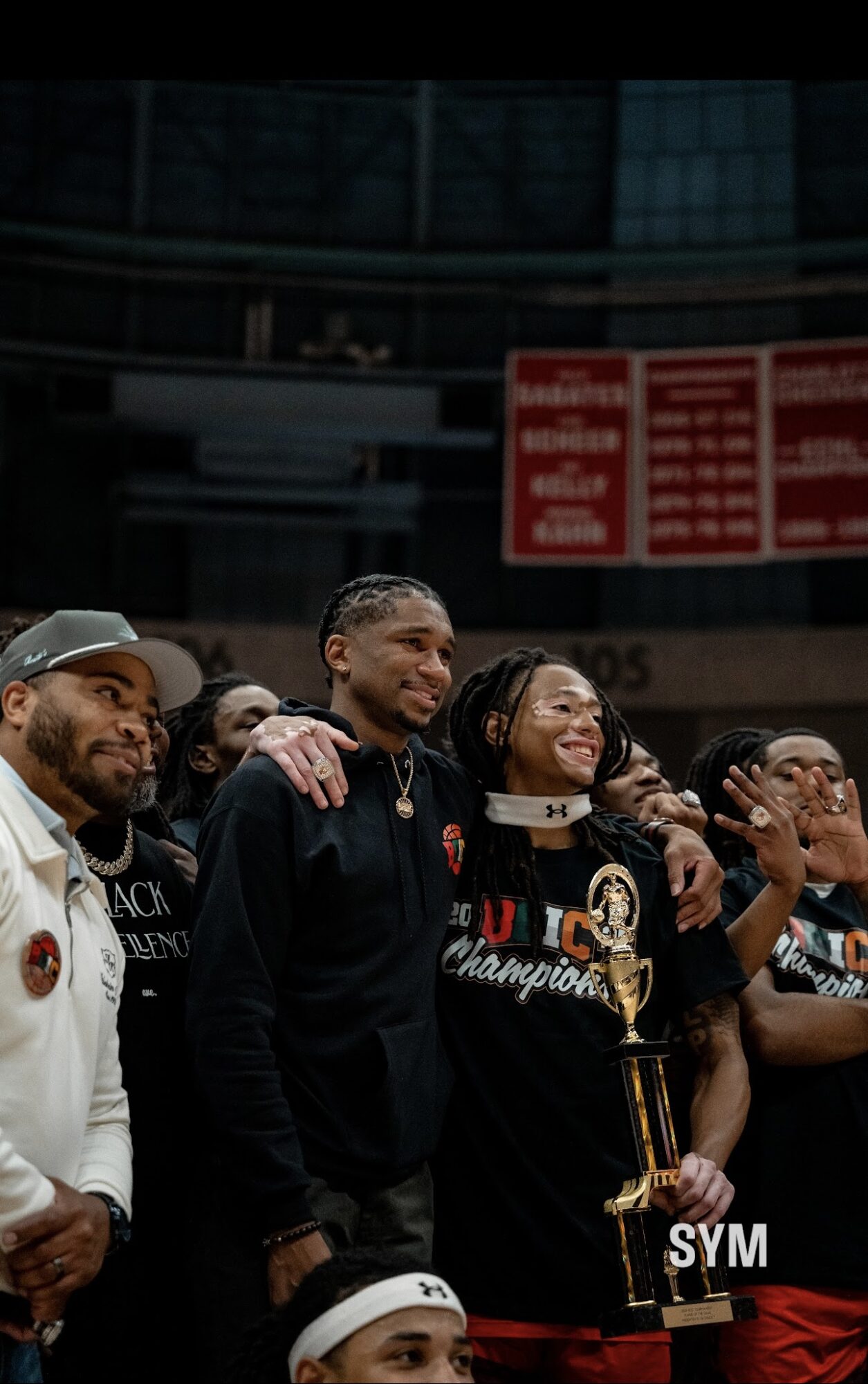 Group of people celebrating, one holding a trophy, smiling, in a sports arena with a scoreboard in the background.