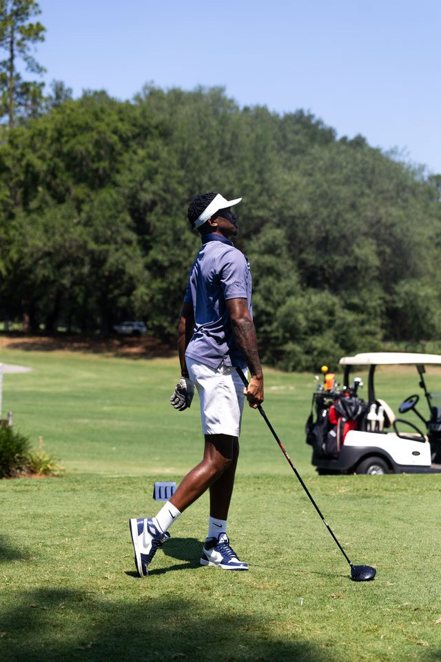 A person in a blue shirt, white shorts, and a white hat stands on a golf course holding a golf club, with trees and a golf cart in the background.