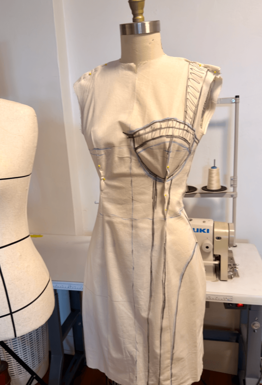 Dress form wearing a beige dress with black pattern lines, in a sewing room with a sewing machine and mannequin.