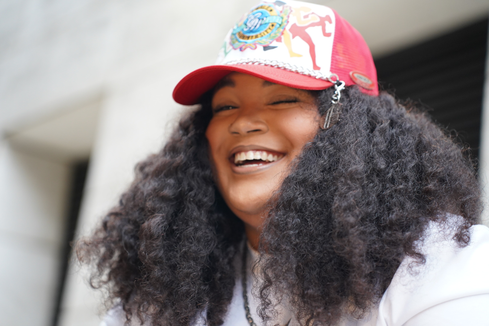 Woman with curly hair smiling, wearing a colorful cap, outdoors, bright lighting.