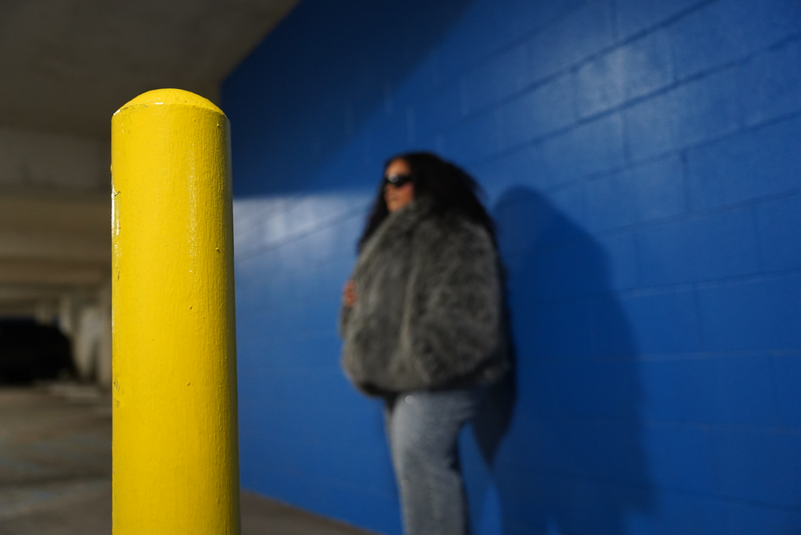 Person with a backpack standing against a blue wall, with a yellow pole in foreground.