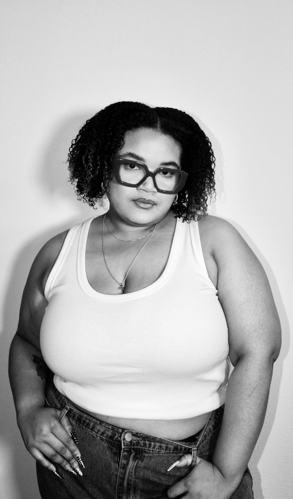 Woman with curly hair, glasses, wearing a white tank top and jeans, standing with hand on hip against a plain background.