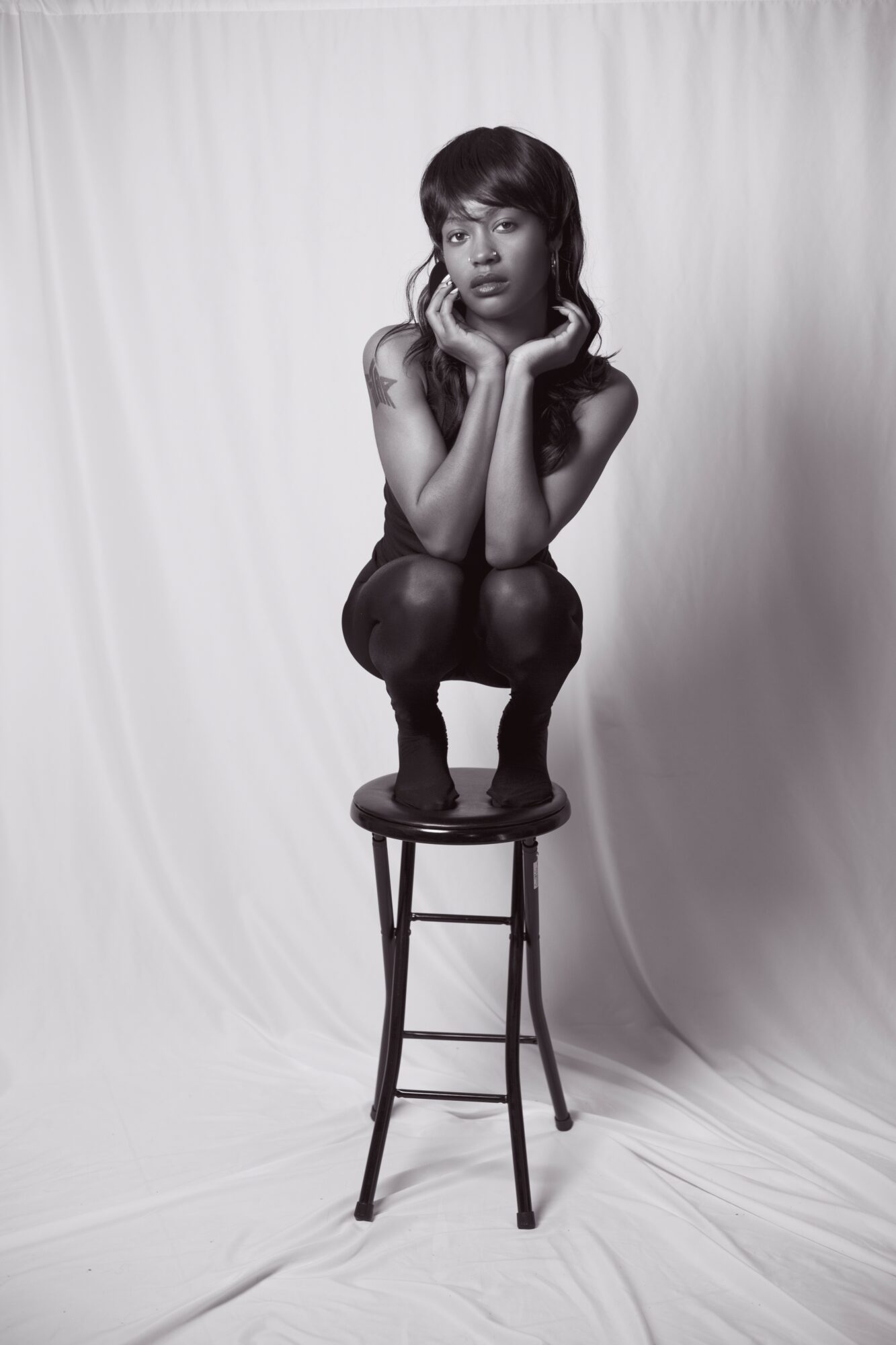 Person with dark hair crouching on a chair, hands on face, against a plain background.