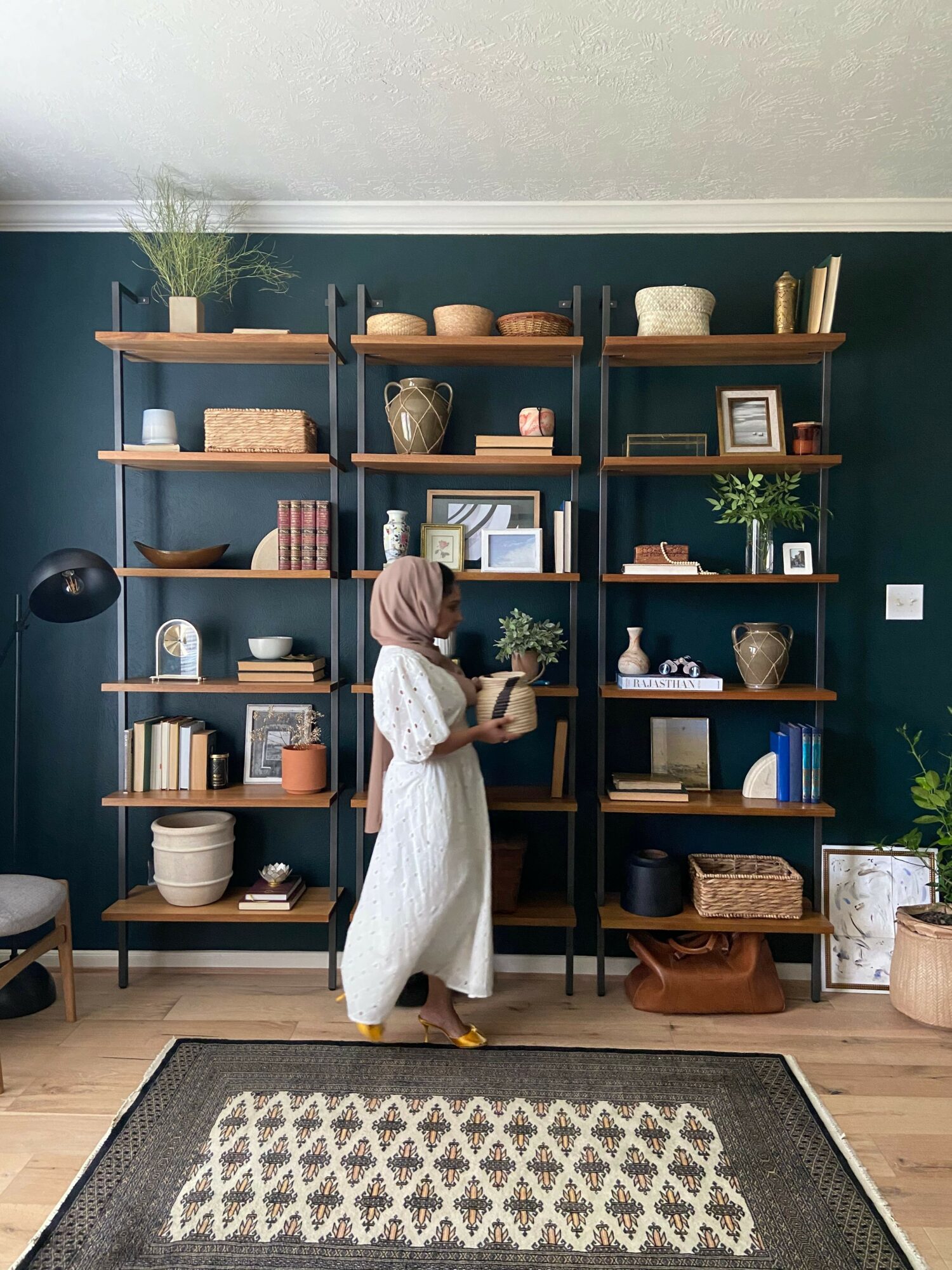 Person in a white dress looking at a bookshelf with decorative items and books, against a dark green wall.