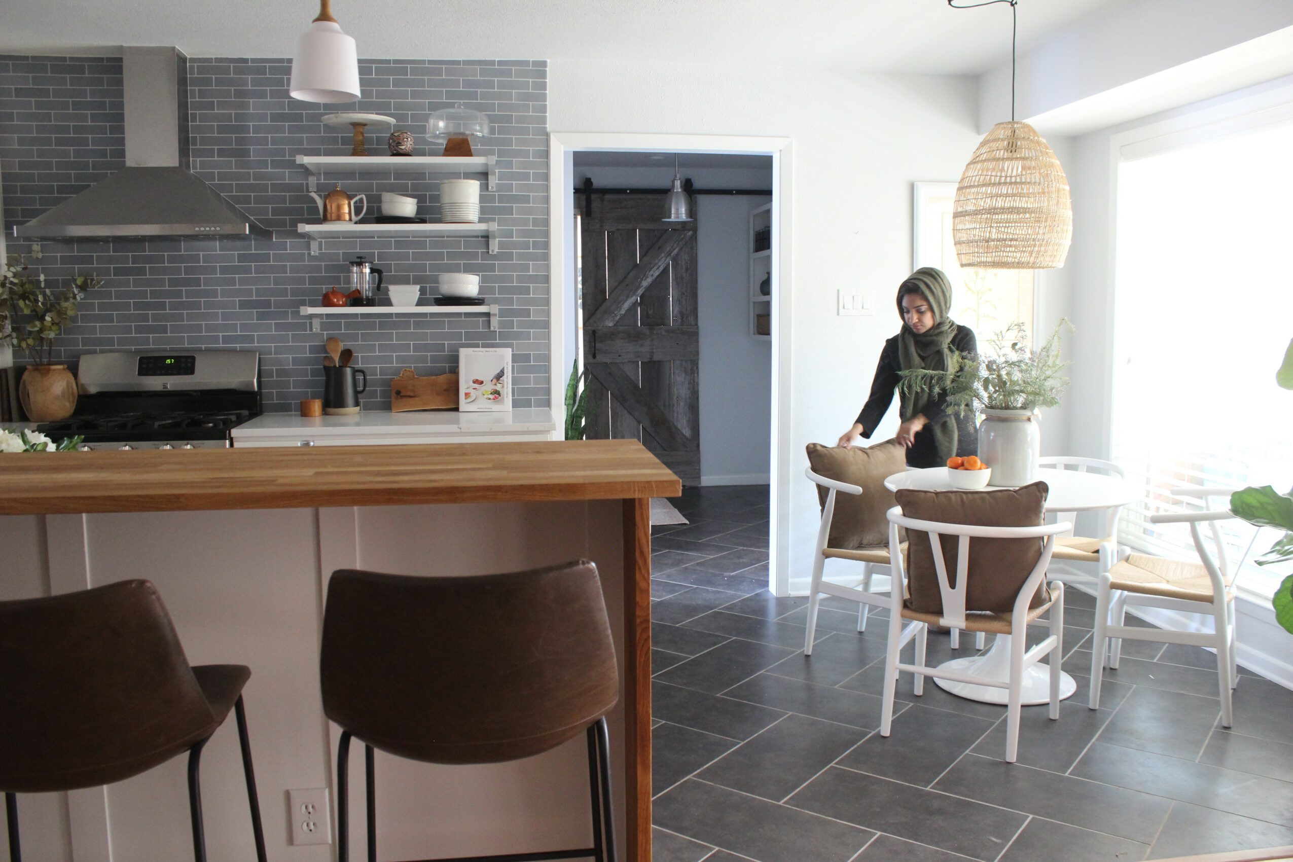 Person standing near a dining table in a modern kitchen and dining area with gray tiled floor and open shelves.