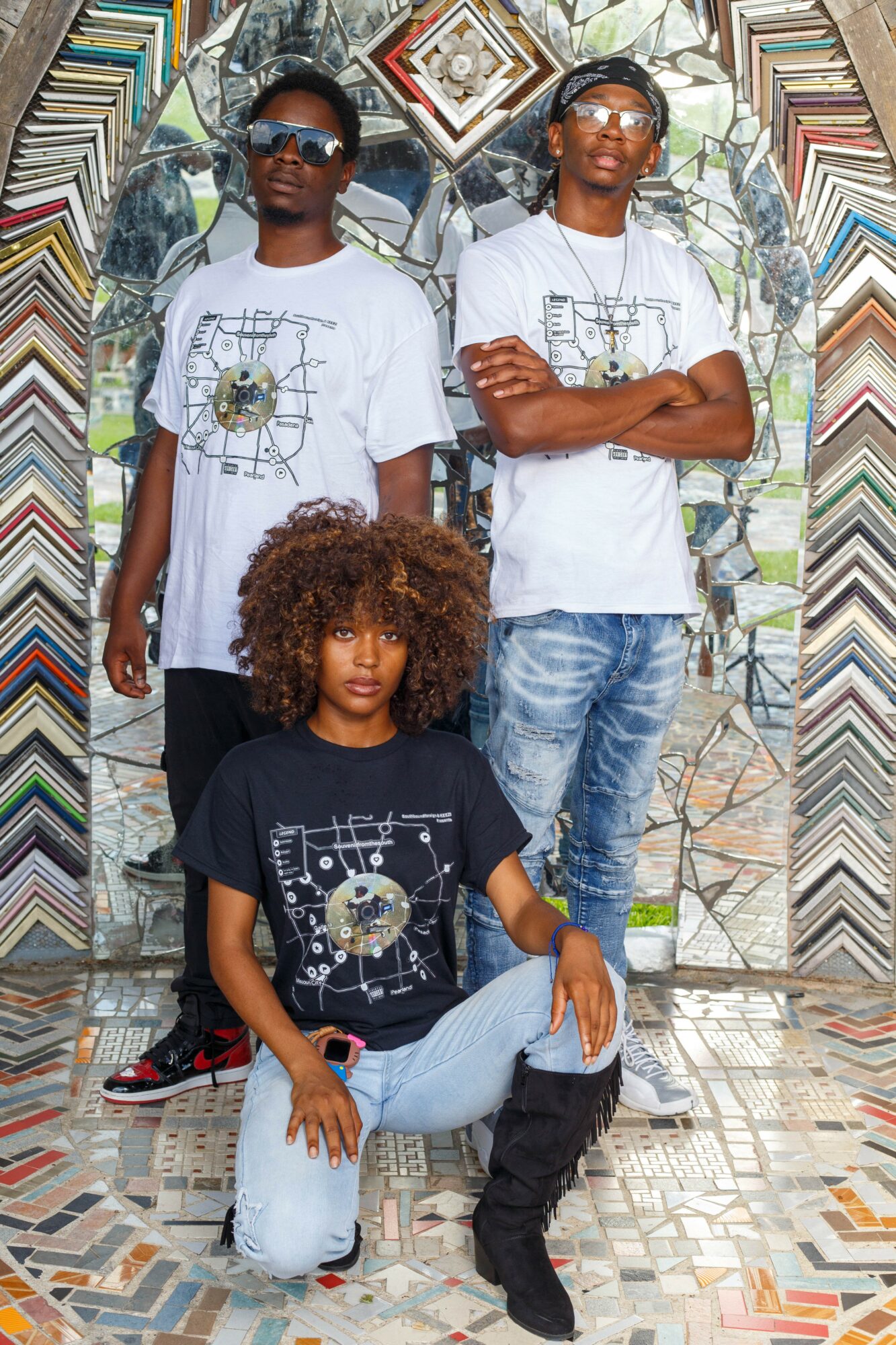 Three people pose outdoors in front of a colorful mosaic wall, two standing and one sitting, all wearing graphic t-shirts.